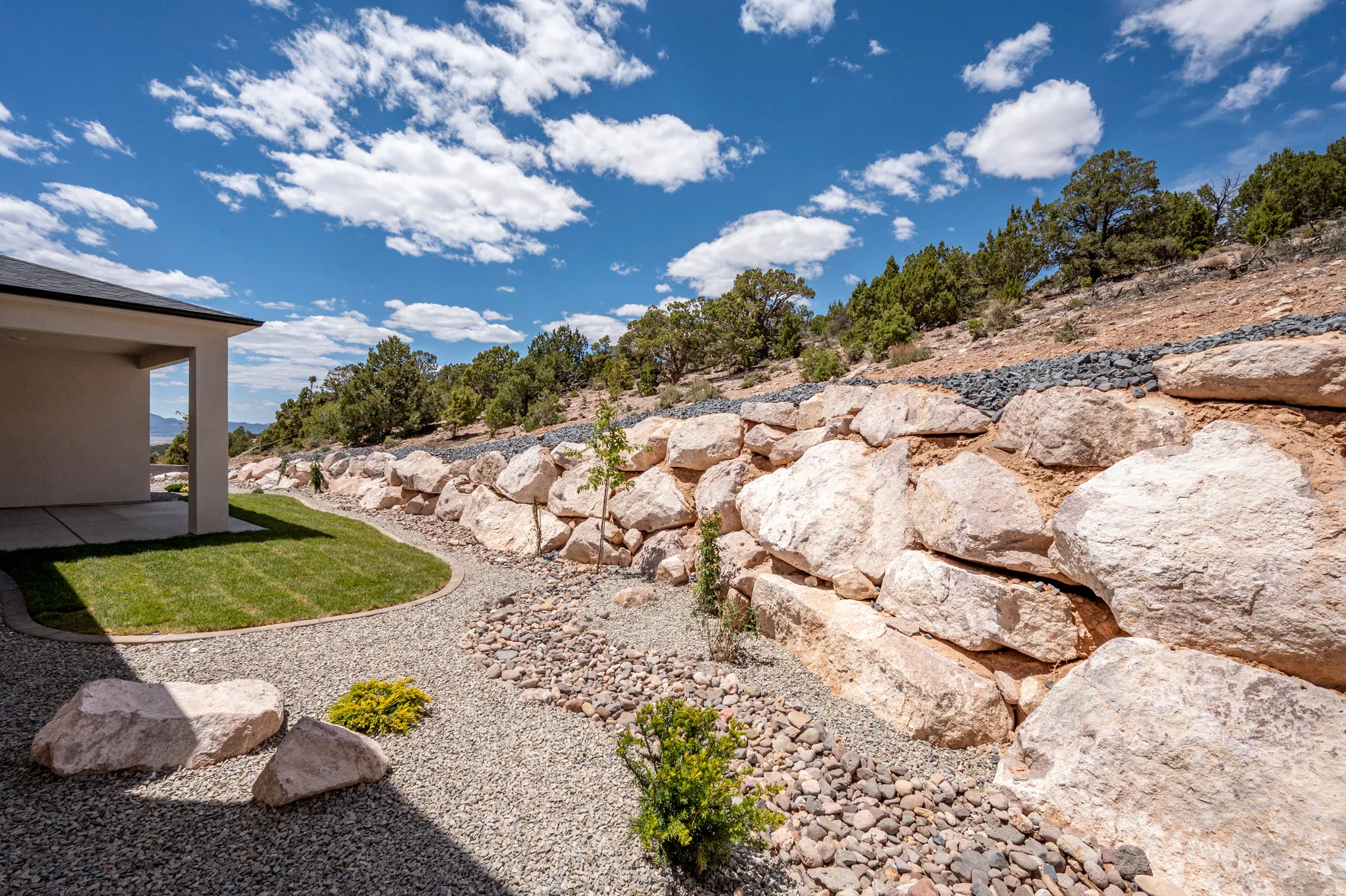 Manicured lawn area with a curved border of decorative gravel, large natural stone retaining wall, and established trees on the hillside