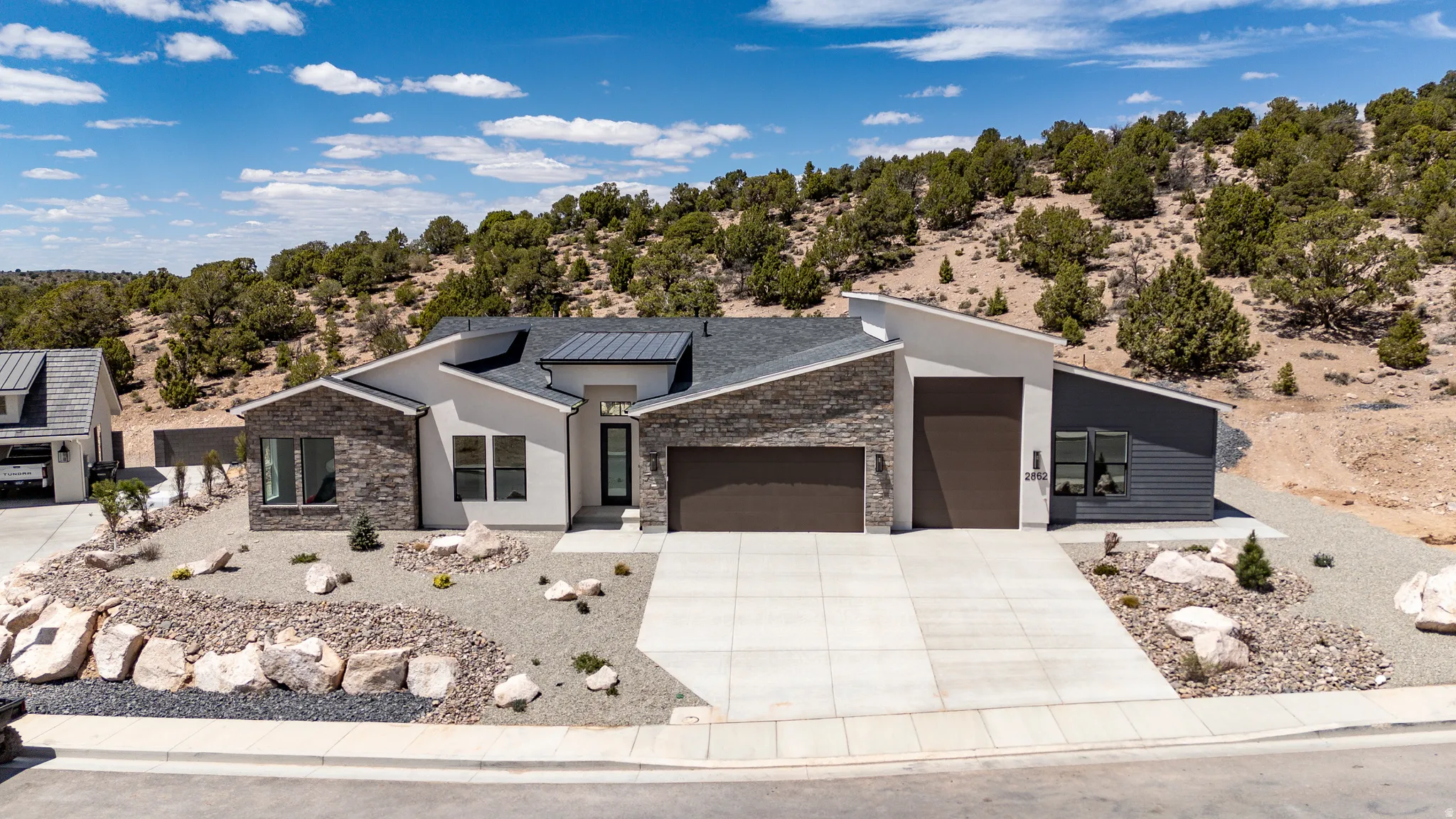 Modern exterior featuring a prominent two-car garage, stone and stucco facade, dark metal roofing, xeriscaped front yard, and an additional single-car garage