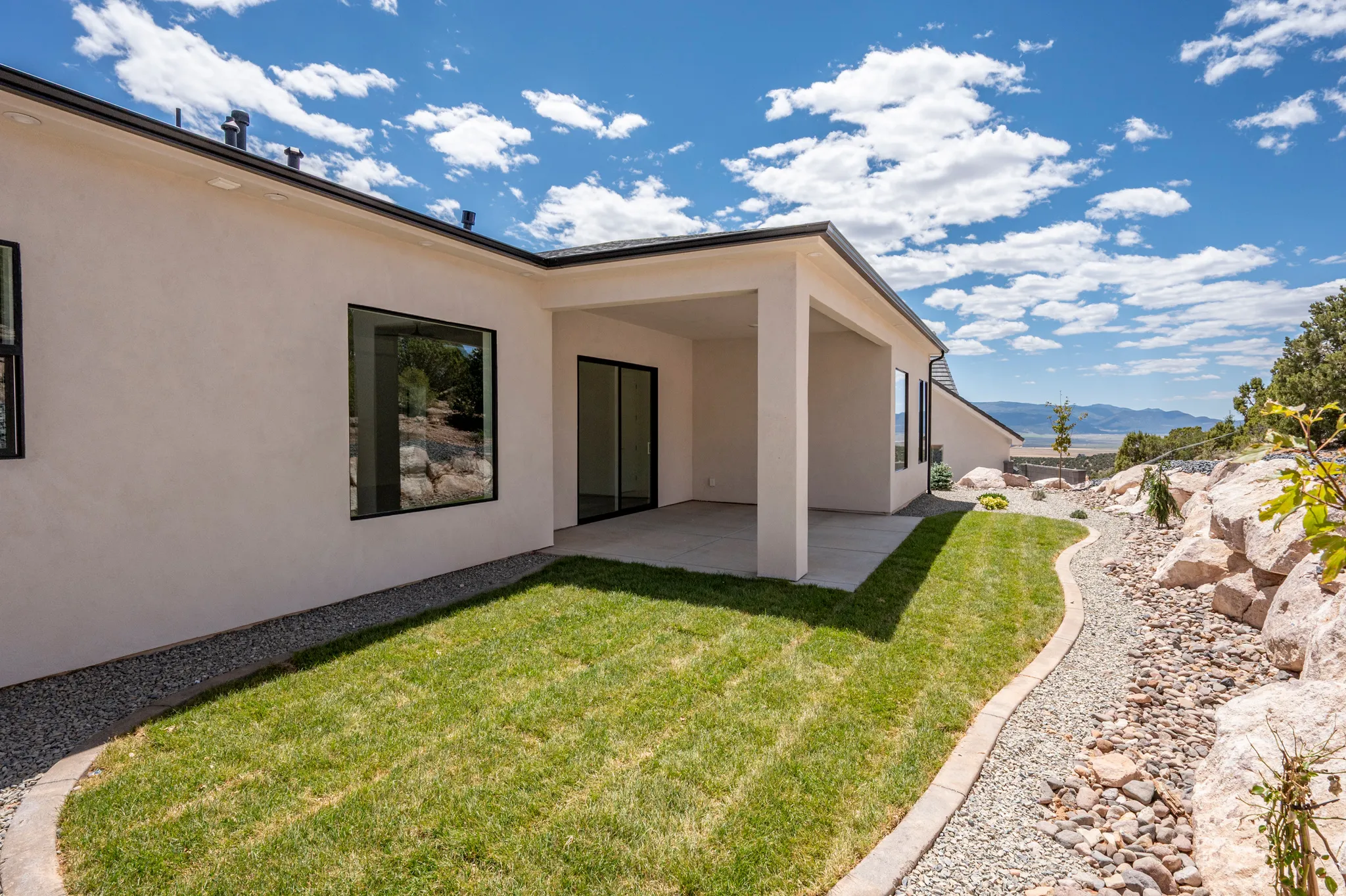 Rear exterior featuring a covered patio, manicured lawn, and a gravel path bordered by natural rock landscaping