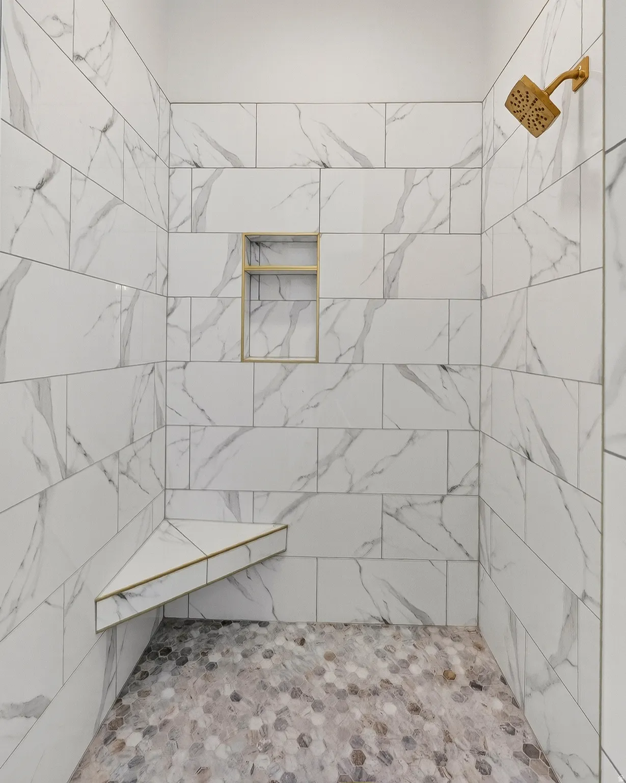 Shower enclosure featuring large format white tile with grey veining, a built-in corner bench, and a recessed niche, all accented with gold trim