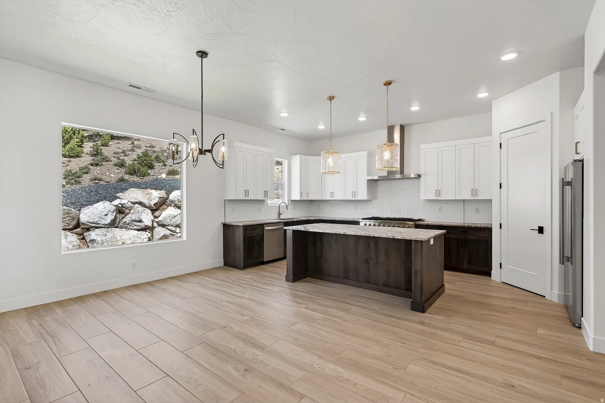 Open-concept kitchen and dining area featuring light wood-finish flooring, a contemporary chandelier, recessed lighting, and a window with natural rock and shrubbery scenery
