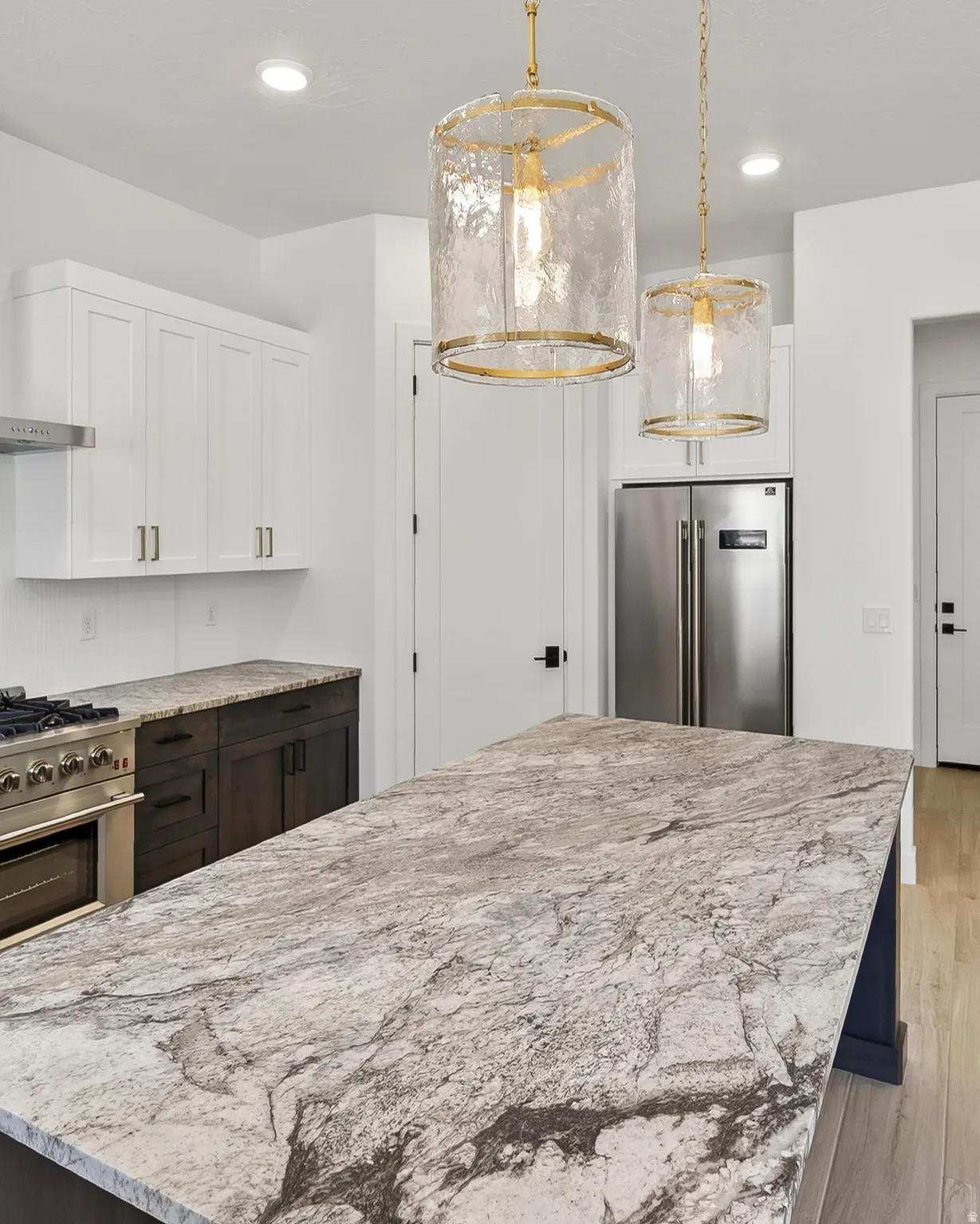 Kitchen island with a patterned stone countertop, two pendant light fixtures, white upper cabinetry, dark lower cabinetry, and stainless steel appliances