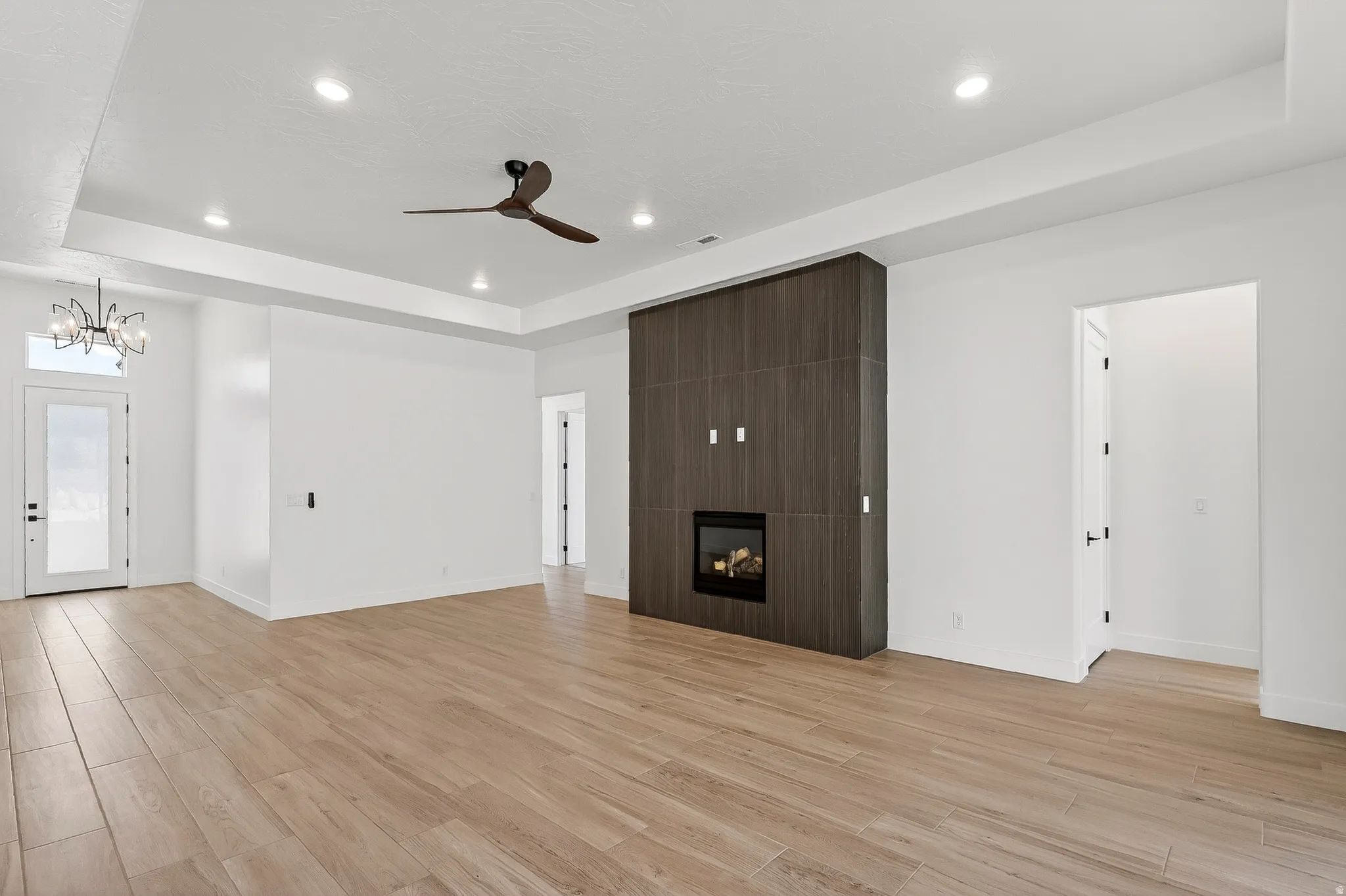Spacious living area featuring wood-finish flooring, a modern fireplace with a dark wood-finish surround, a contemporary ceiling fan, recessed lighting, and a tray ceiling