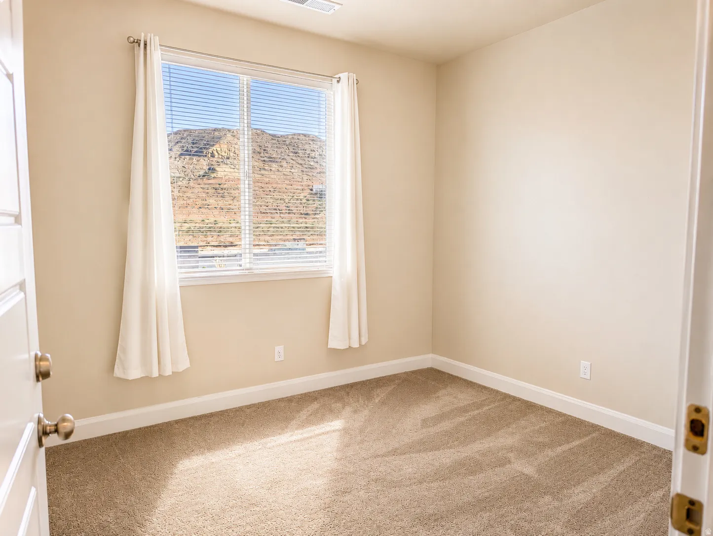 Neutral-toned carpeted room featuring a large window with white blinds and drapes
