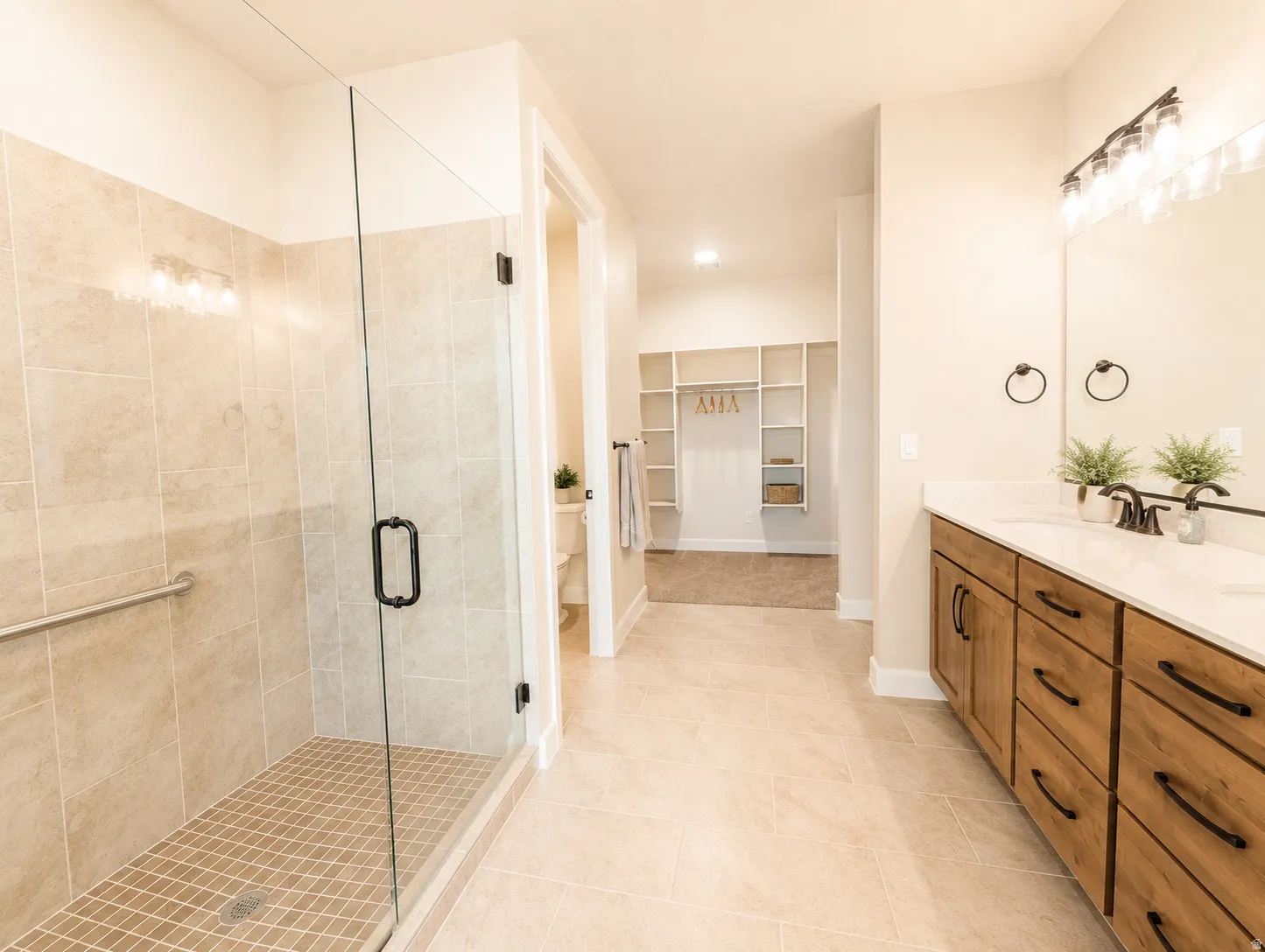 Spacious bathroom featuring a glass-enclosed shower with tile surround, double vanity with wood-finish cabinetry, light stone-finish countertops, and tile flooring