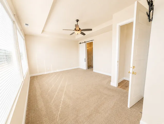 Spacious room featuring neutral carpeting, a tray ceiling with a dark-tone ceiling fan, and multiple windows with blinds