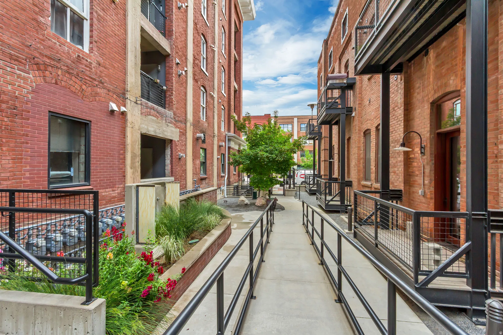 Concrete walkway with black metal railings framed by brick facades featuring individual balconies with black metal railings, and integrated landscaping with mature trees and vibrant foliage