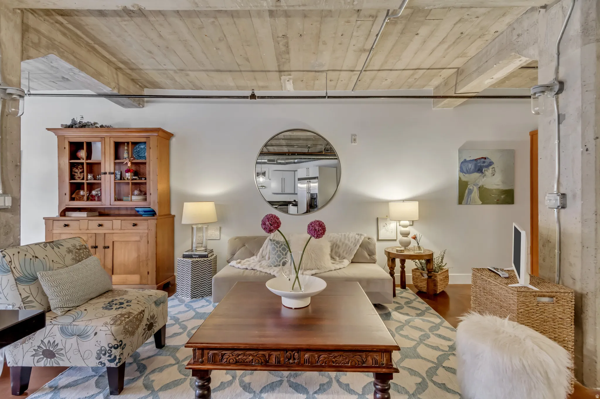 Spacious room featuring exposed concrete ceilings with visible wood beams, industrial-style conduit lighting, and light hardwood flooring partially covered by an area rug
