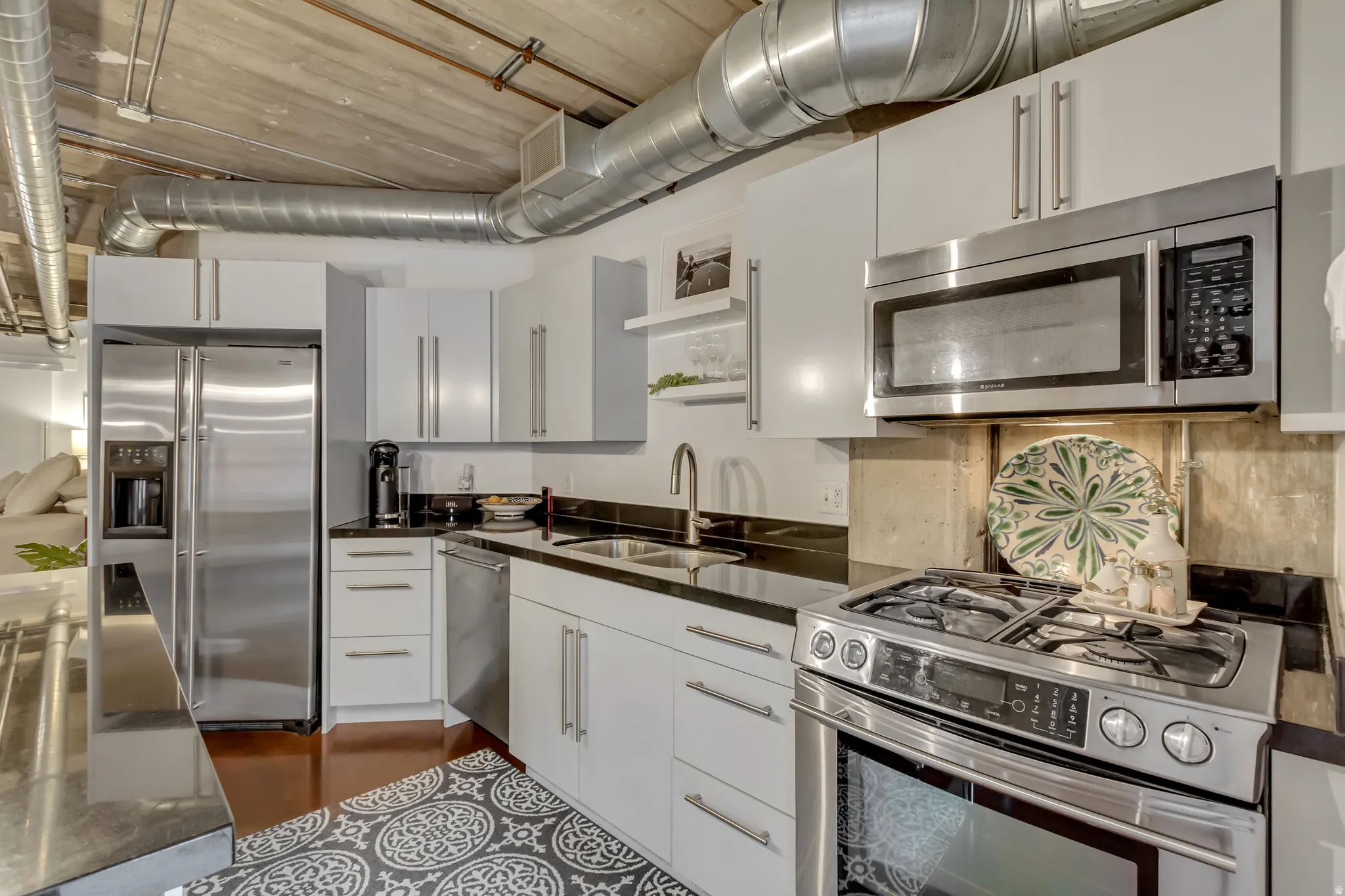 Modern kitchen featuring stainless steel appliances, dark countertops, white cabinetry, exposed ductwork, and warm-toned flooring