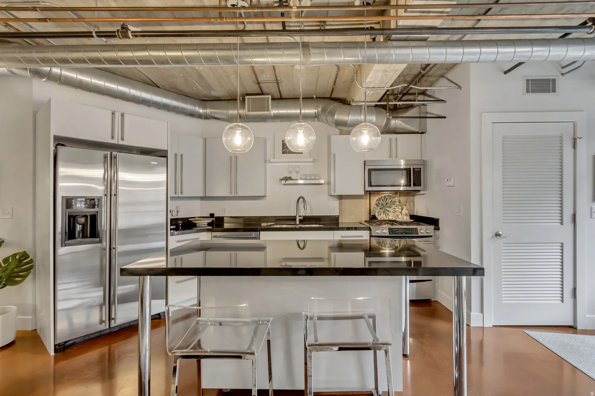 Industrial-inspired kitchen featuring an exposed ceiling with ductwork, stainless steel appliances, a kitchen island with a dark countertop, white cabinetry, and globe pendant lighting