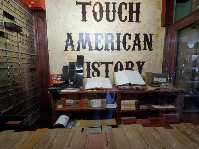 Vintage bank interior featuring a prominent "TOUCH AMERICAN HISTORY" wall graphic, a wooden display counter with shelving, a large wall of safe deposit boxes, and a glass-fronted cabinet