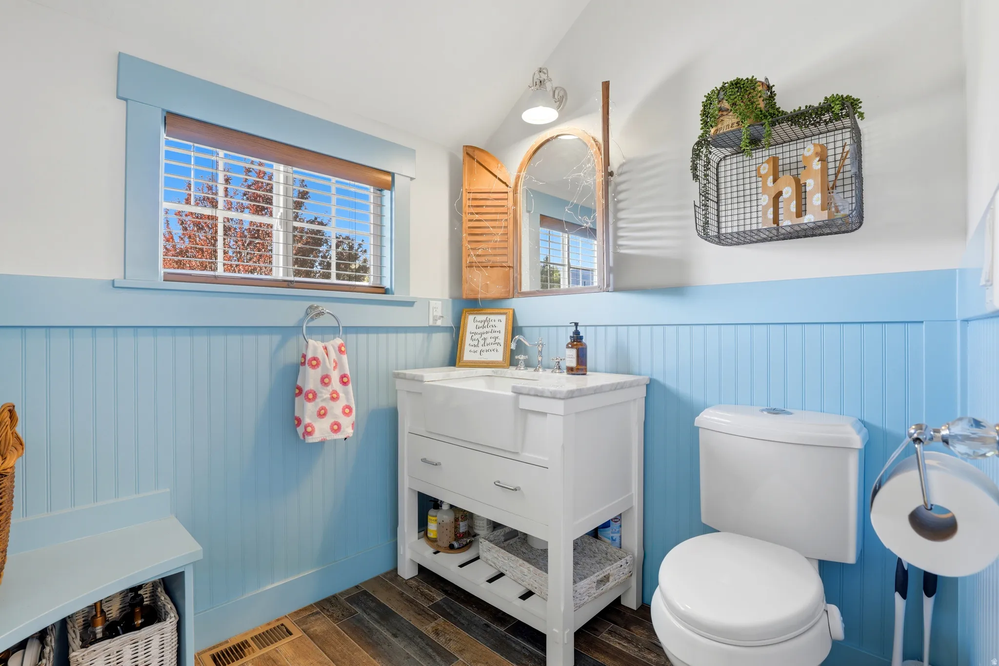 Bathroom featuring a white vanity with an integrated sink and countertop, wood-finish flooring, a window with horizontal blinds, beadboard wainscoting, and a white toilet