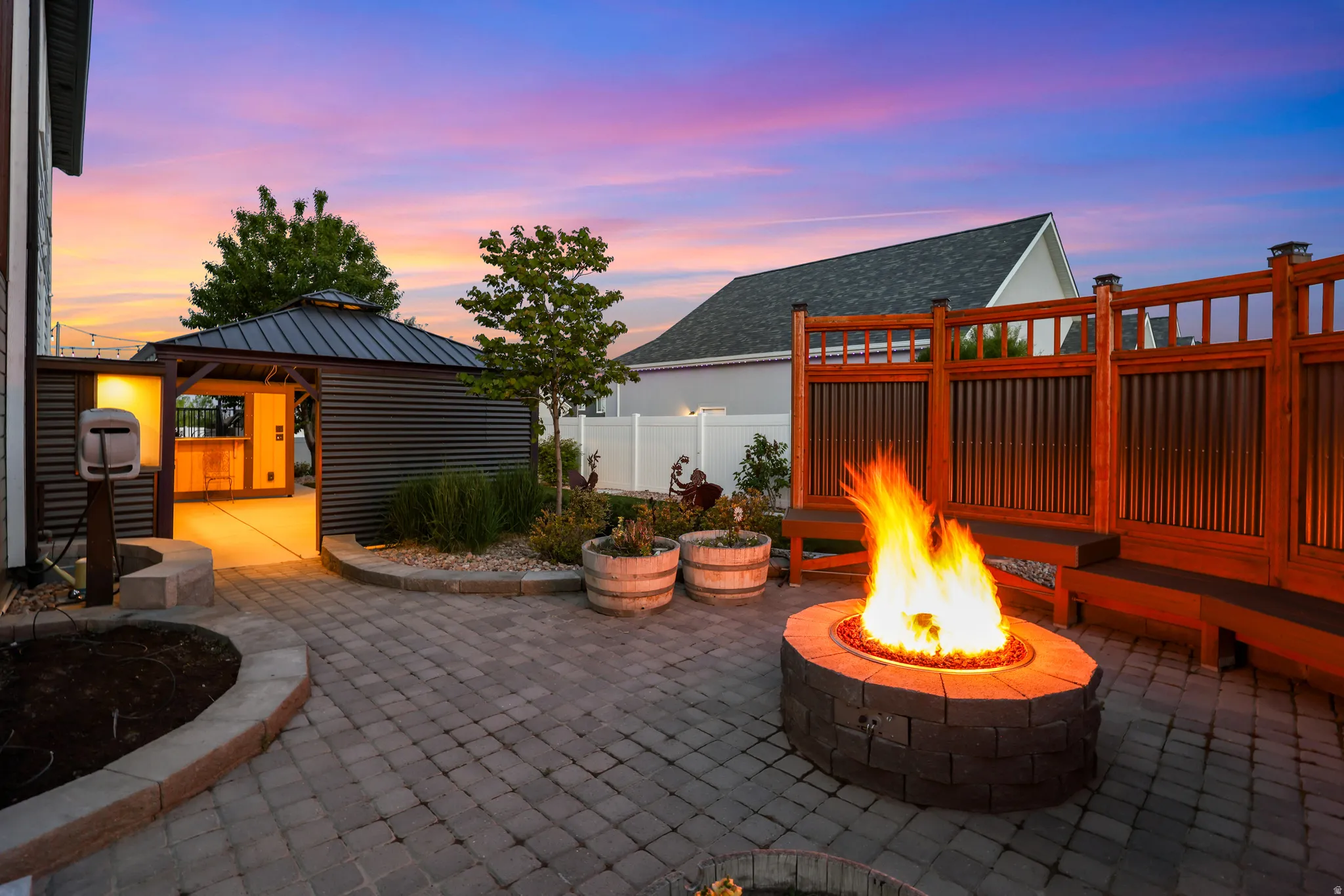 Paver patio featuring a stone fire pit, built-in wooden bench seating, and a corrugated metal gazebo with an integrated bar
