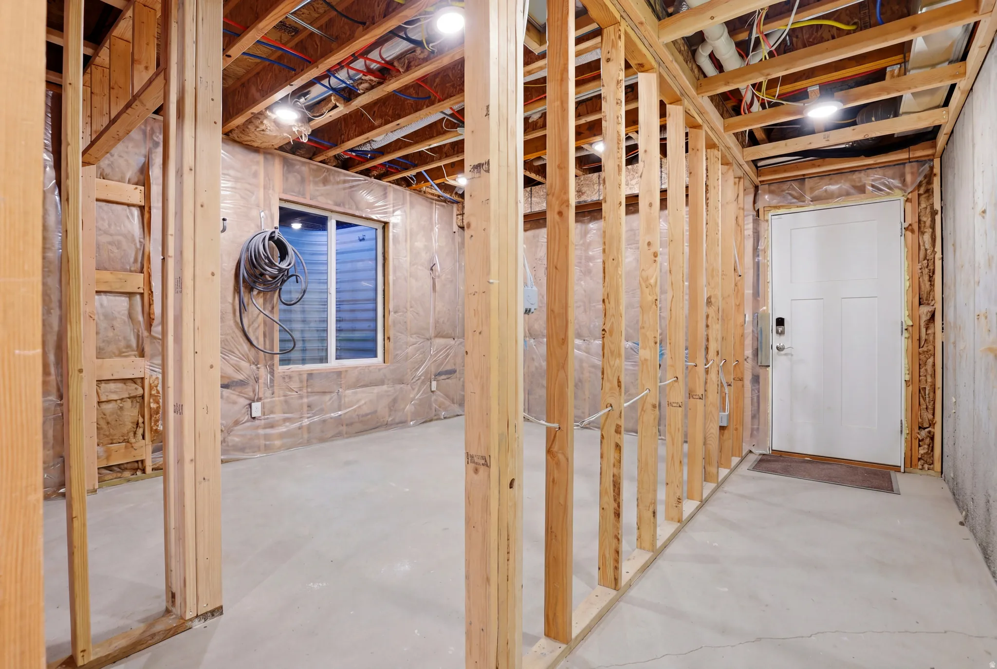 Framed interior space with a concrete floor, exposed ceiling joists with wiring and plumbing, and a white paneled door