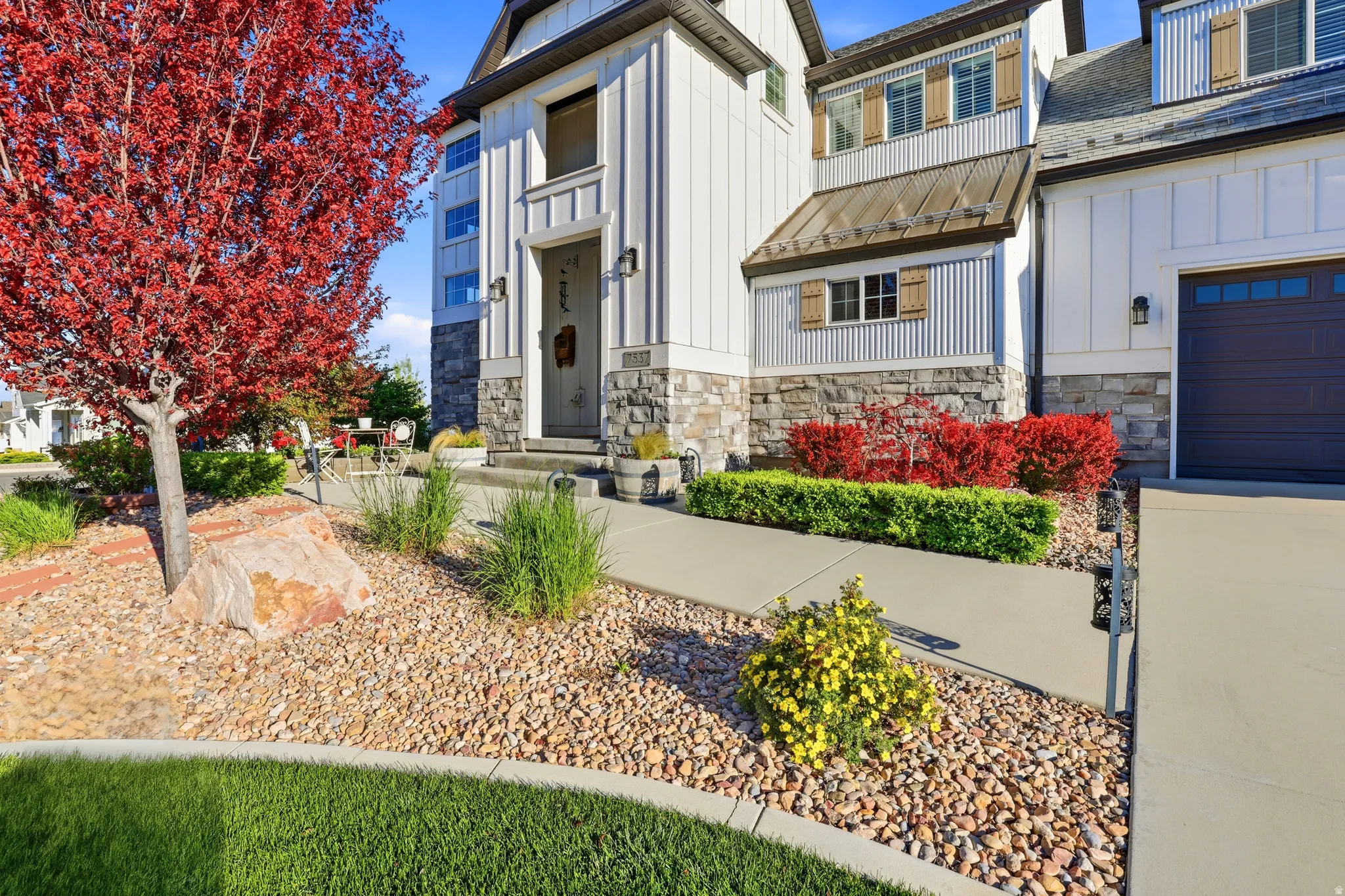 Modern farmhouse exterior with vertical white siding, stone accents, a dark garage door, a corrugated metal awning, and decorative wooden shutters