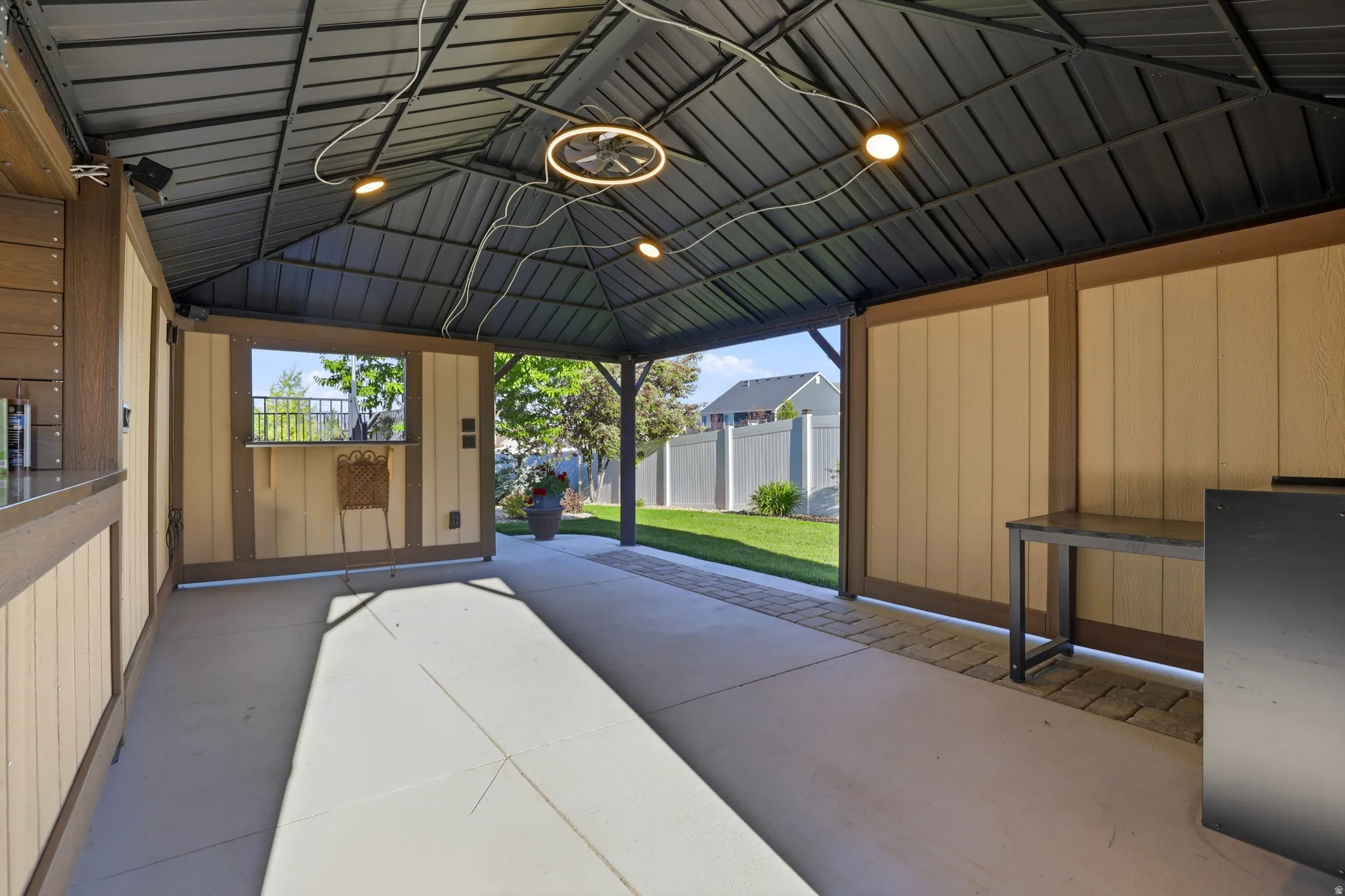 Expansive covered patio featuring a metal gazebo roof with integrated lighting, a built-in bar with a serving window, concrete flooring, and a paver stone border