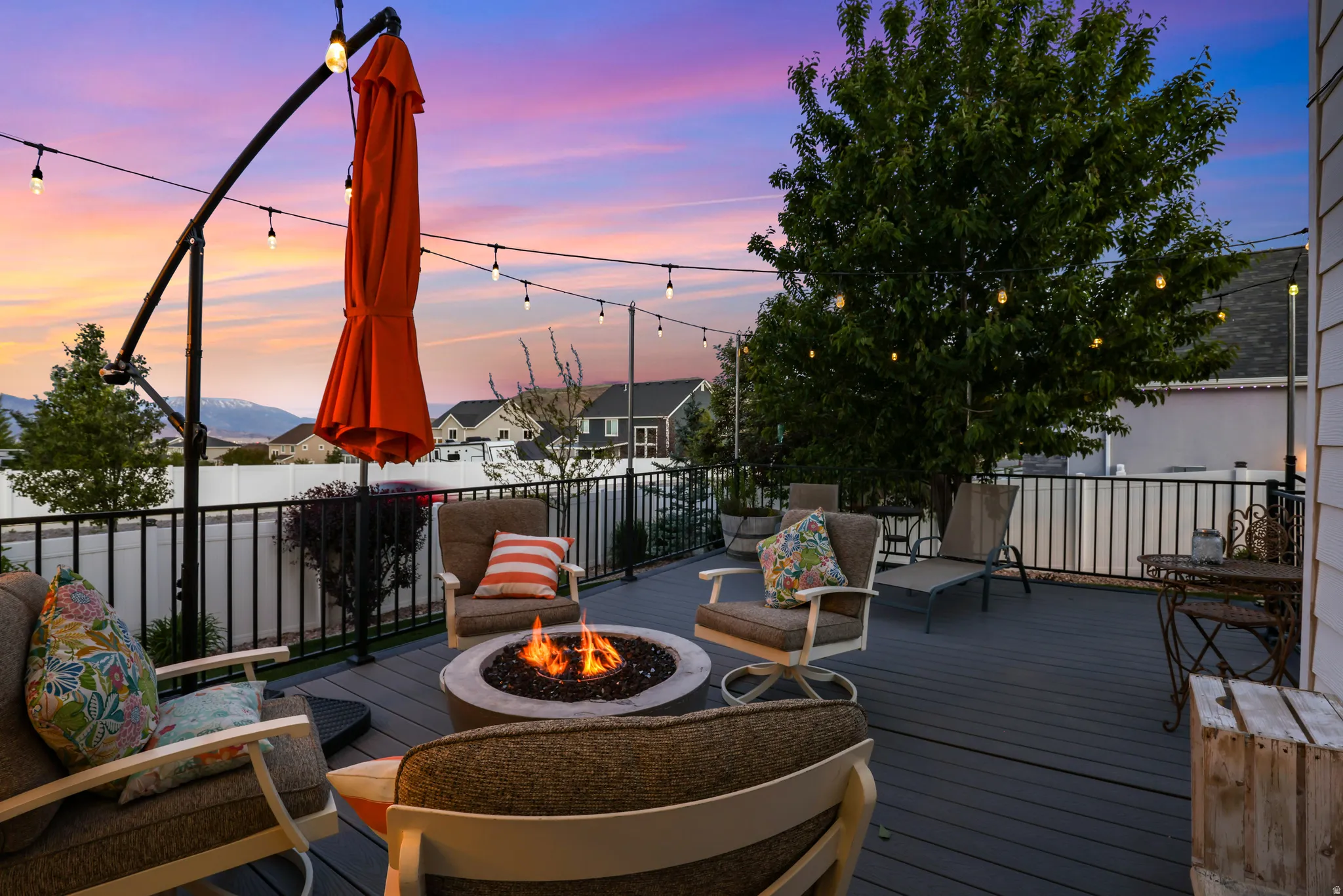 Spacious elevated deck featuring composite decking, a built-in fire pit, black metal railings, string lights, and mature tree landscaping