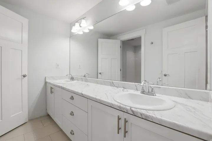 Spacious vanity featuring double undermount sinks, a faux-marble countertop, white cabinetry with silver hardware, and a large wall-mounted mirror