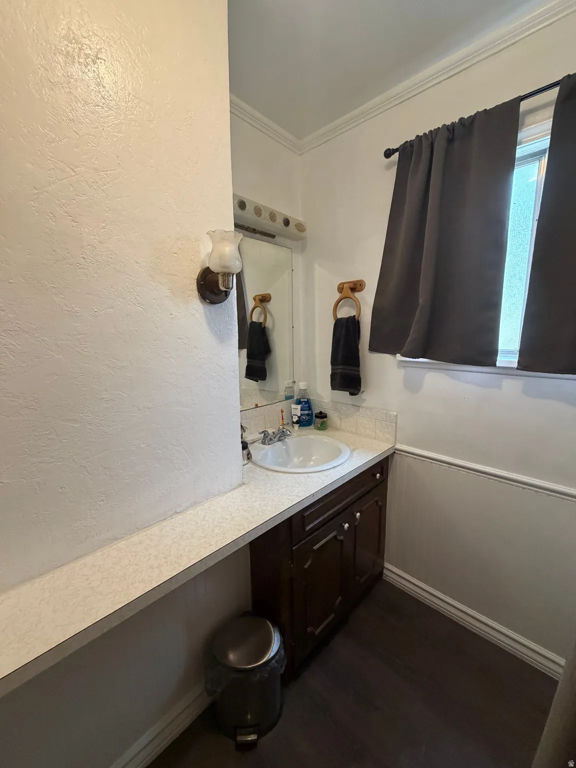 Bathroom vanity featuring a white countertop with integrated sink, dark wood-finish cabinetry, a wall-mounted light fixture, and crown molding