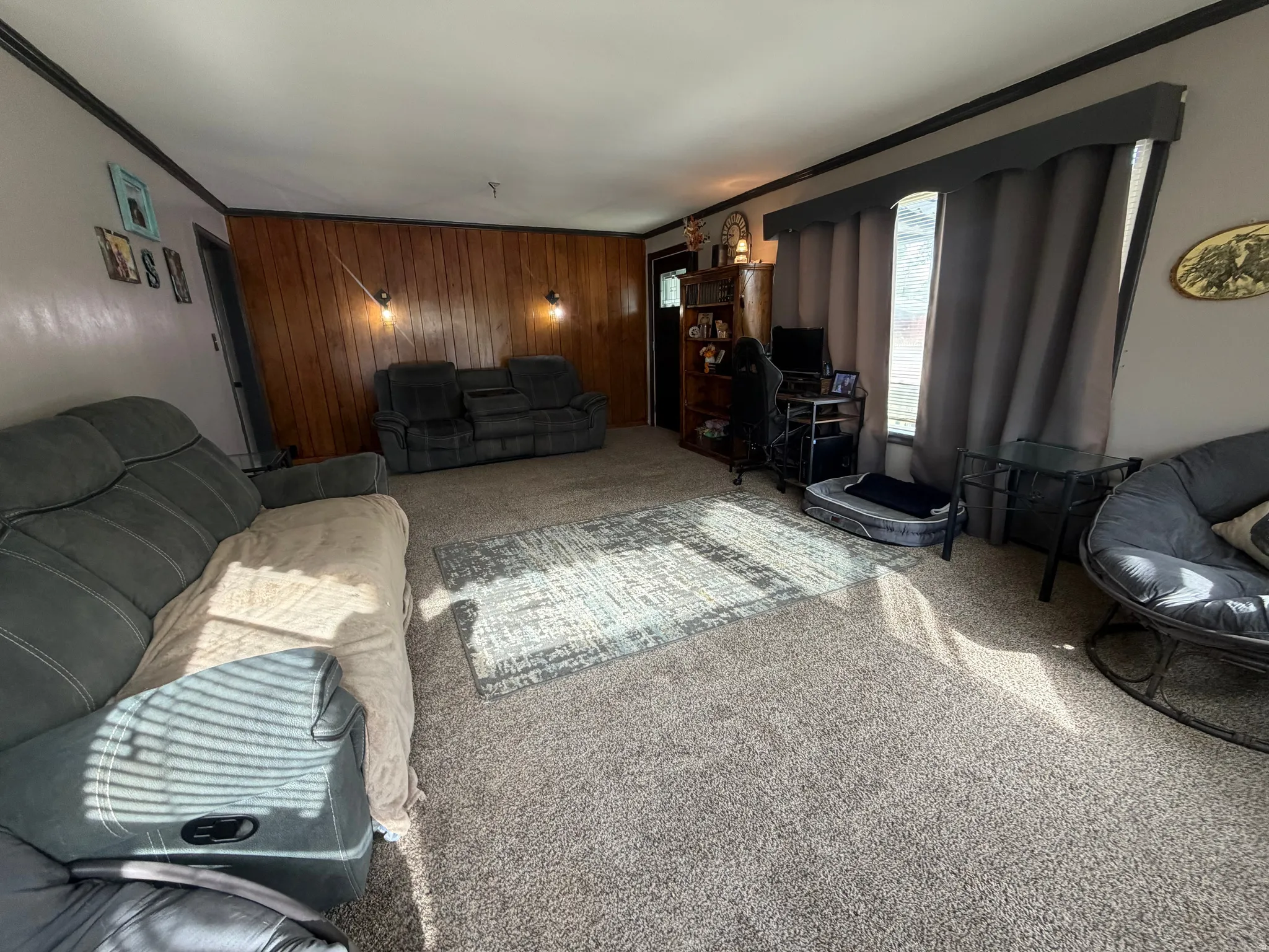 Living area featuring wood-finish accent wall with sconce lighting, light-colored carpet flooring, and a window with valance and drapes