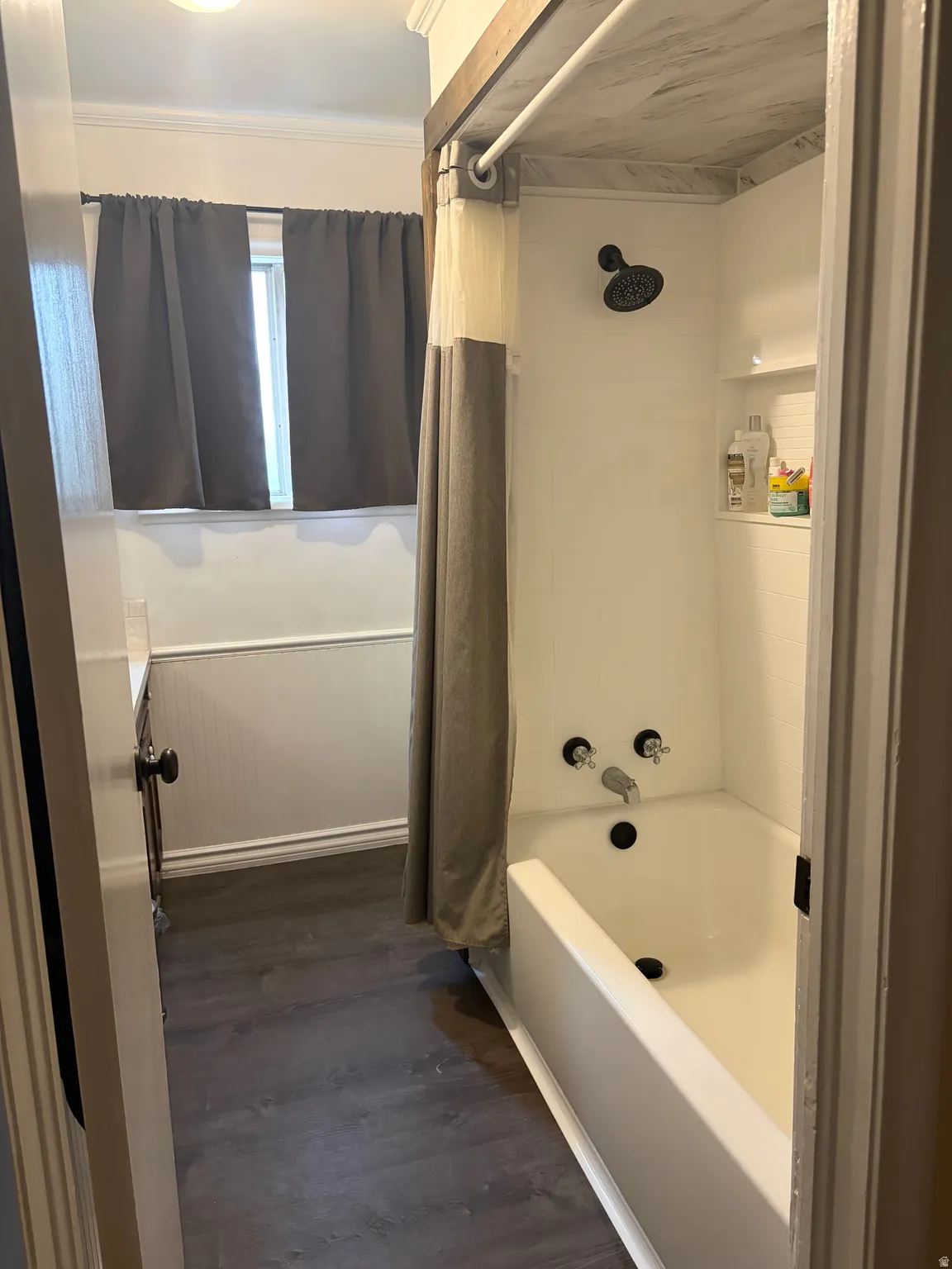 Bathroom featuring a shower-tub combination with black fixtures, dark wood-finish flooring, white beadboard wainscoting, and a window with crown molding