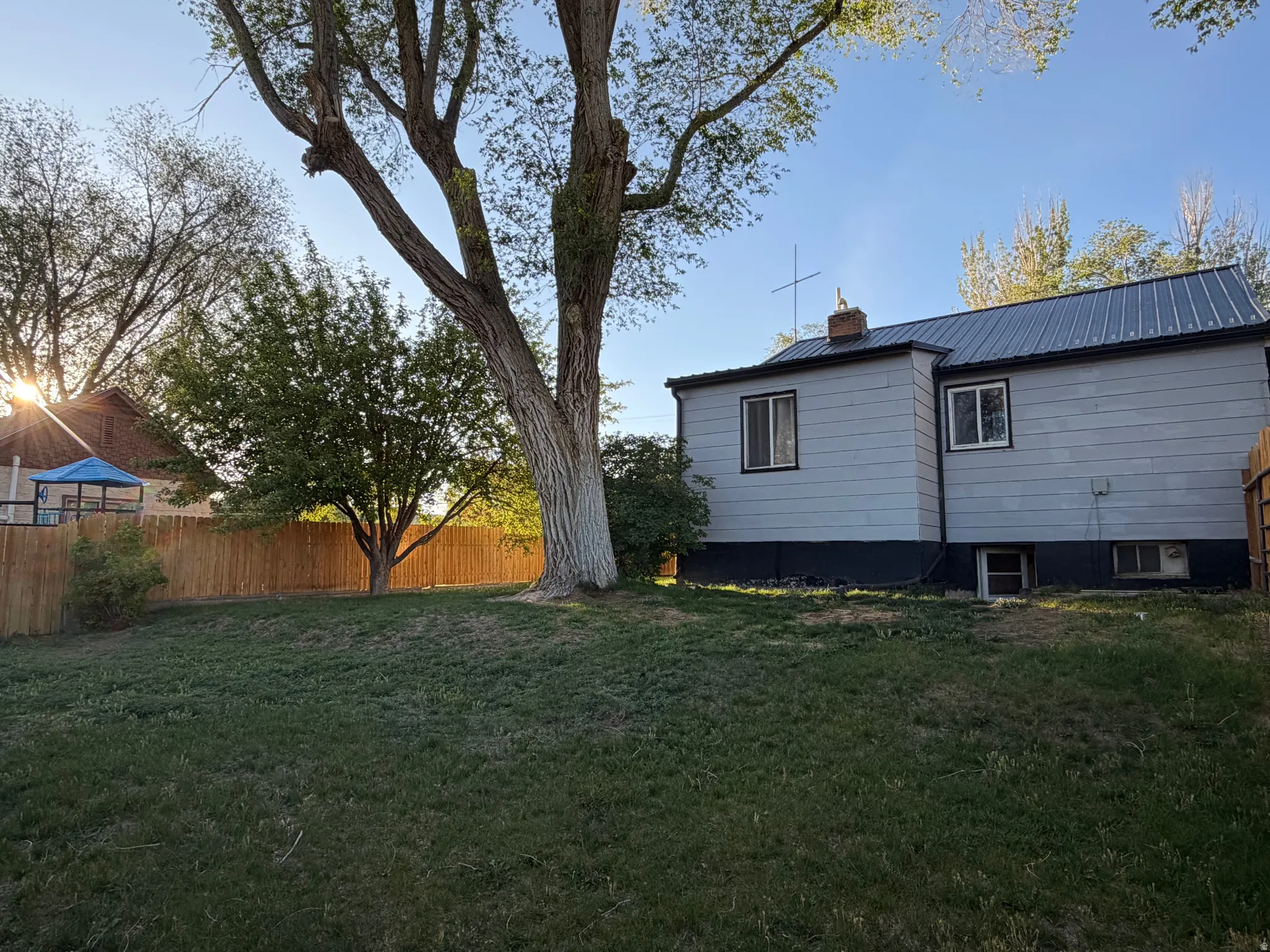 Expansive rear yard featuring mature trees, a privacy fence, and a light-colored exterior with a dark metal roof