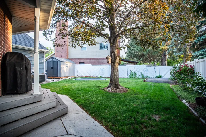 Expansive backyard featuring a manicured lawn, mature tree, white privacy fencing, storage shed, and a composite deck