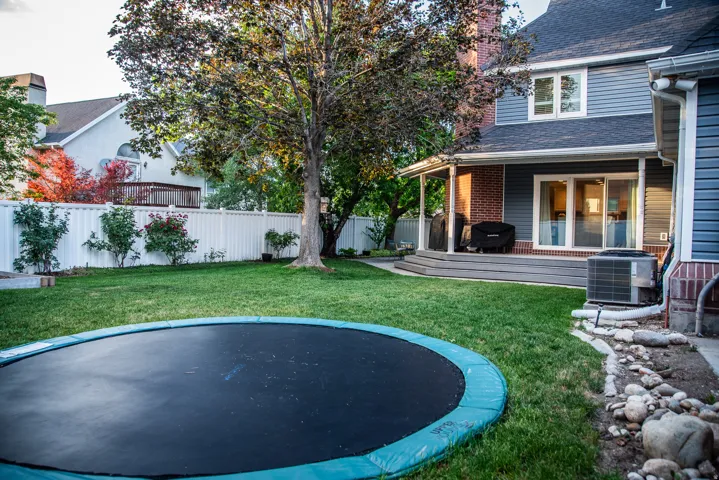 Spacious backyard featuring a manicured lawn, mature trees, a white privacy fence, and a covered porch with composite decking.  Included in-ground trampoline.