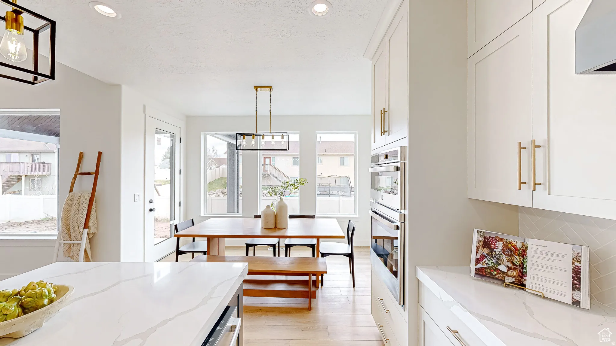 Modern kitchen and dining area featuring light wood-finish flooring, white cabinetry with gold-tone hardware, and integrated stainless steel double ovens