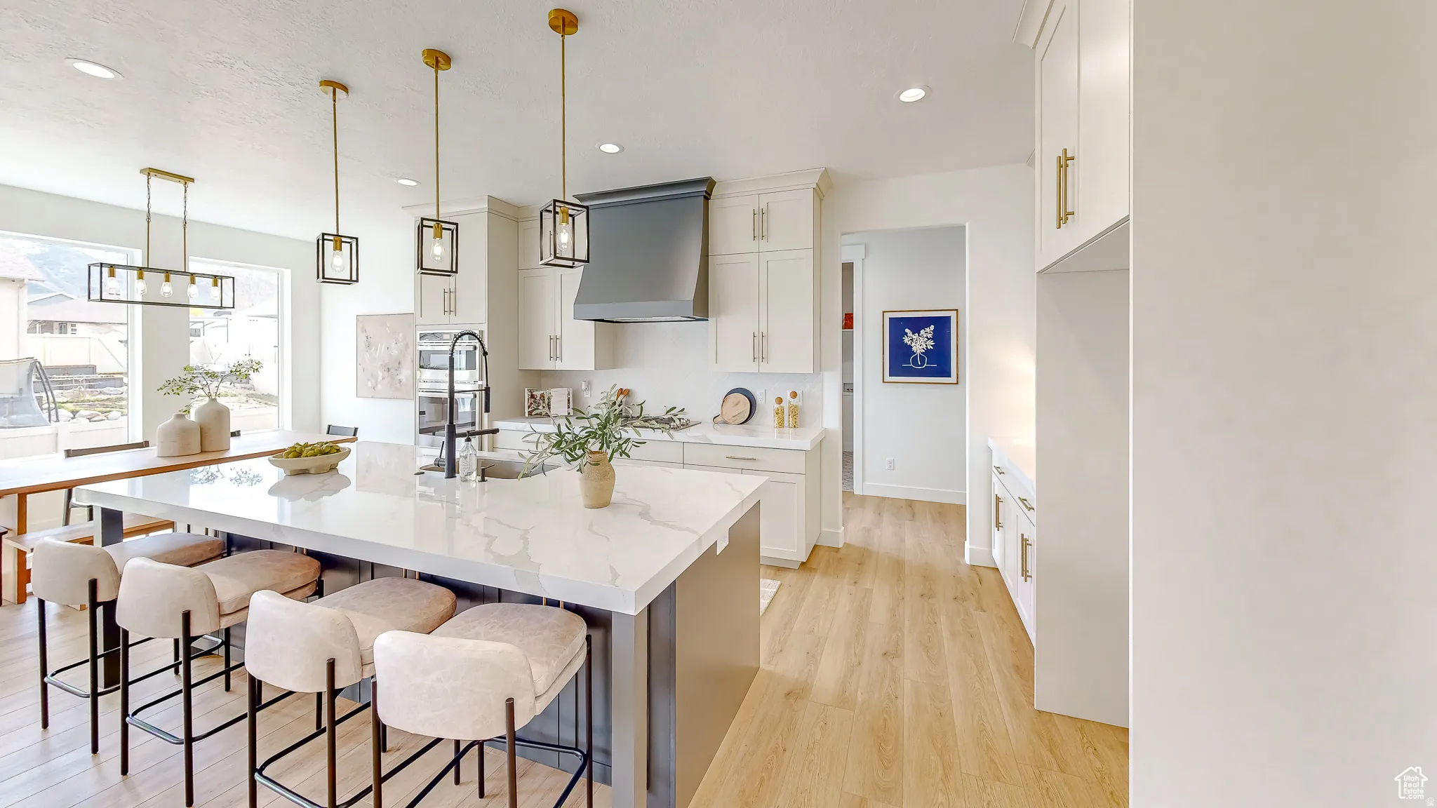 Spacious kitchen featuring an expansive island with a marble-look countertop, white shaker cabinetry, gold-tone hardware, wood-finish flooring, and contemporary pendant lighting