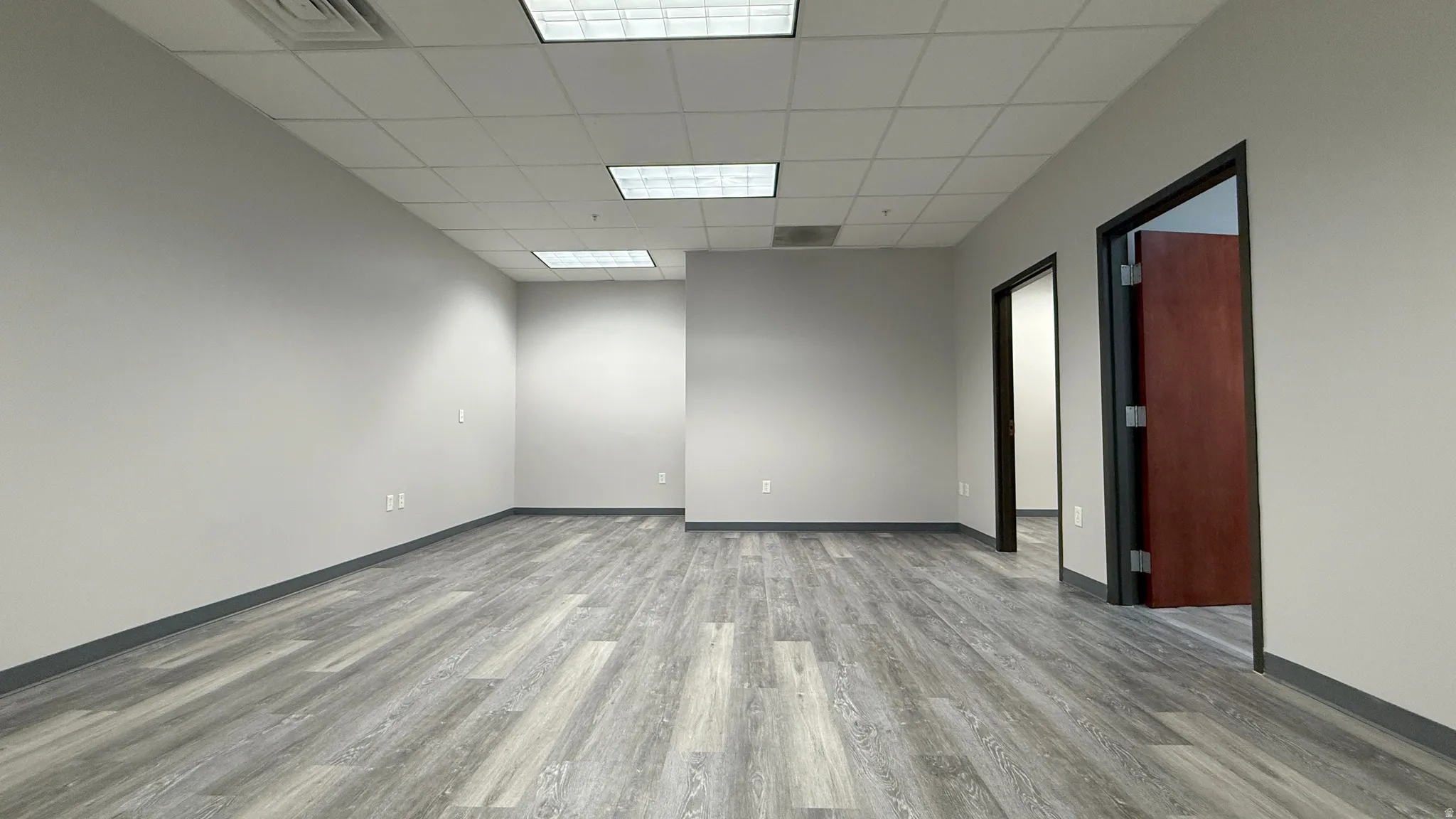 Spacious interior room featuring wood-finish flooring, light gray walls, a dropped ceiling with recessed lighting, and a dark brown door with dark trim