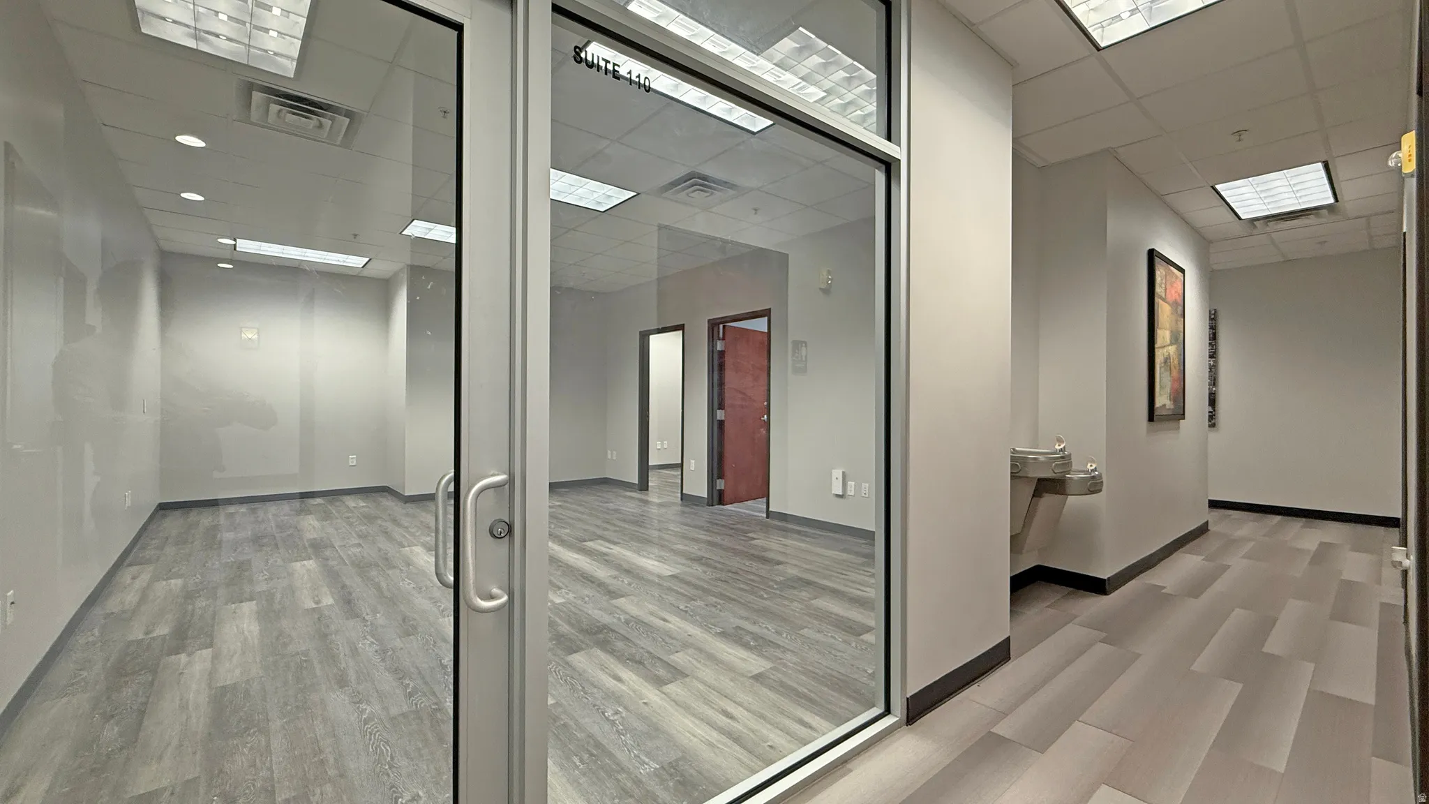 Office suite entrance with glass doors, showcasing wood-finish flooring, recessed lighting, and a drop ceiling