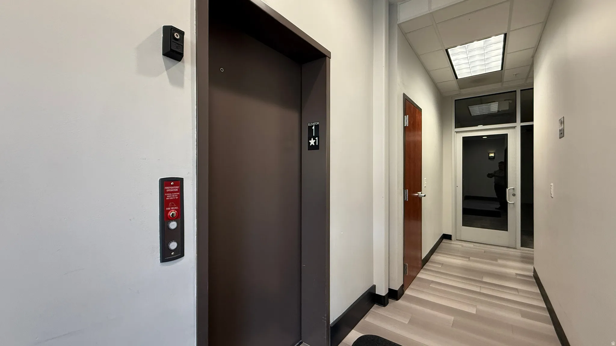 Hallway featuring an elevator with a brushed metal door, light wood-finish flooring, a suspended acoustic tile ceiling with recessed lighting, a paneled wood door with a lever handle, and a glass-paneled entry door with transom