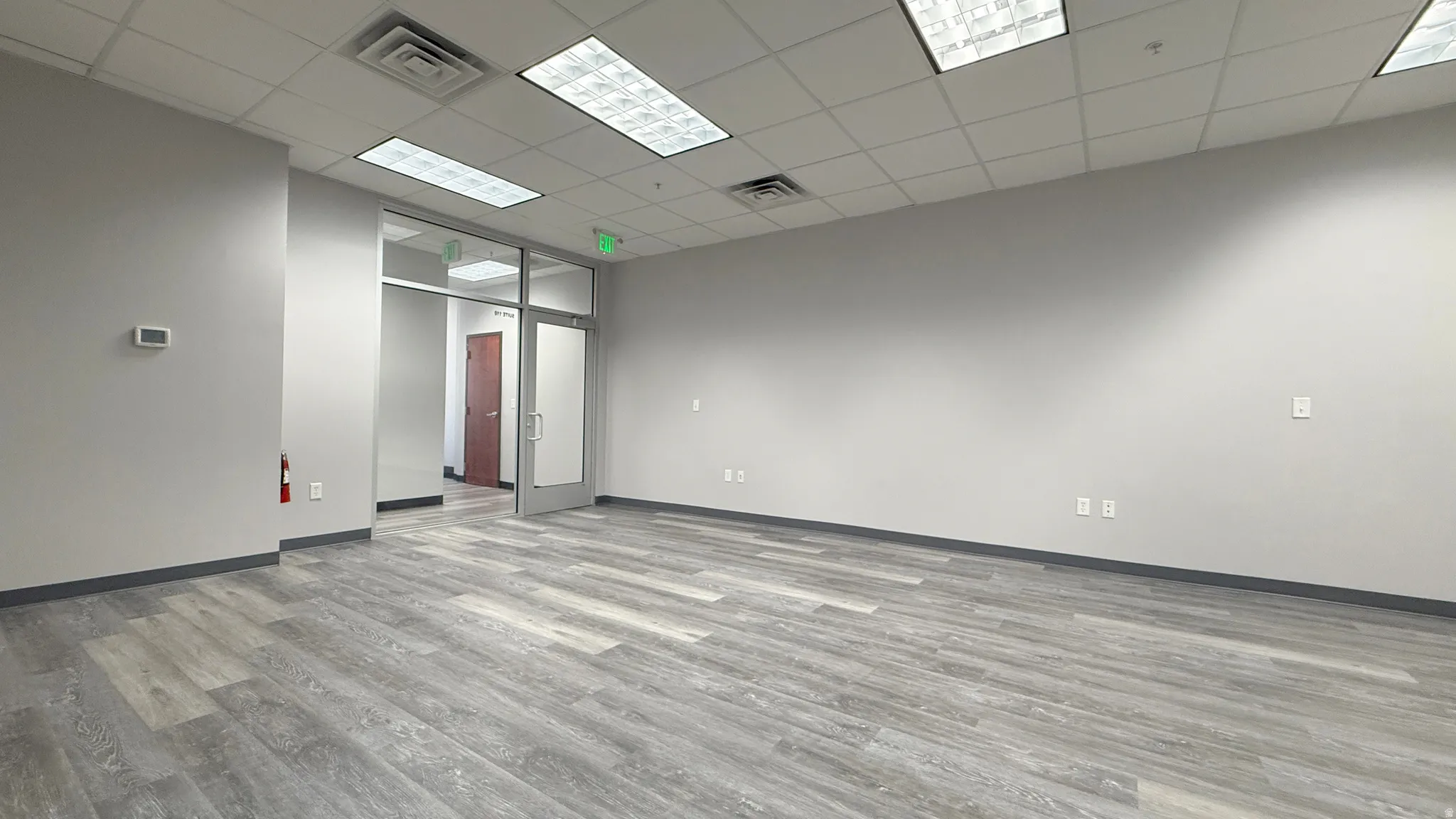 Spacious open-concept commercial interior featuring wood-finish flooring, drop ceiling with integrated fluorescent lighting, light gray painted walls, and a glass-paneled entry door