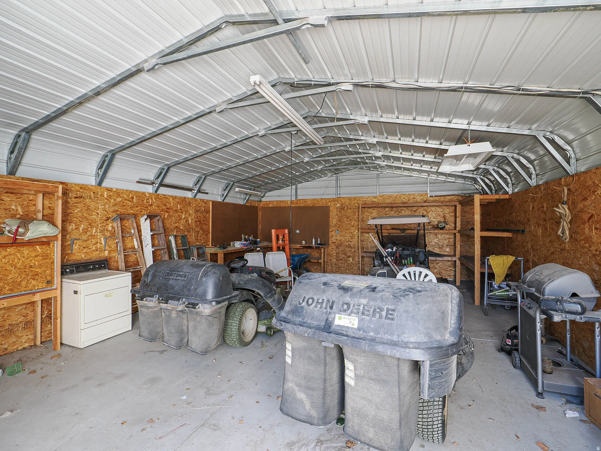 Metal carport structure featuring a corrugated metal roof, exposed metal framing, oriented strand board walls, built-in wooden shelving, and concrete flooring