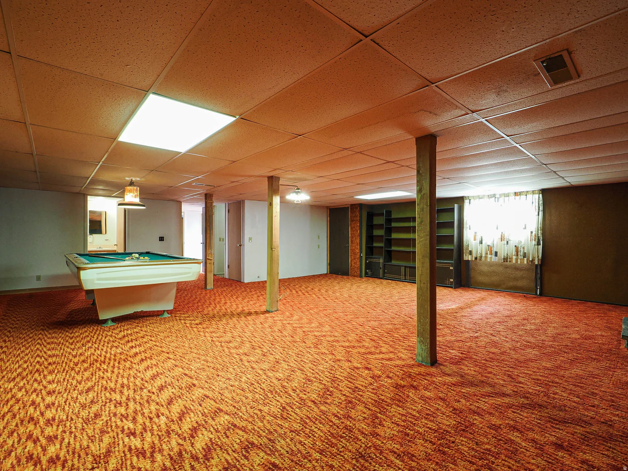 Spacious lower-level area featuring patterned carpeting, drop ceiling with recessed lighting, exposed structural beams, and built-in shelving