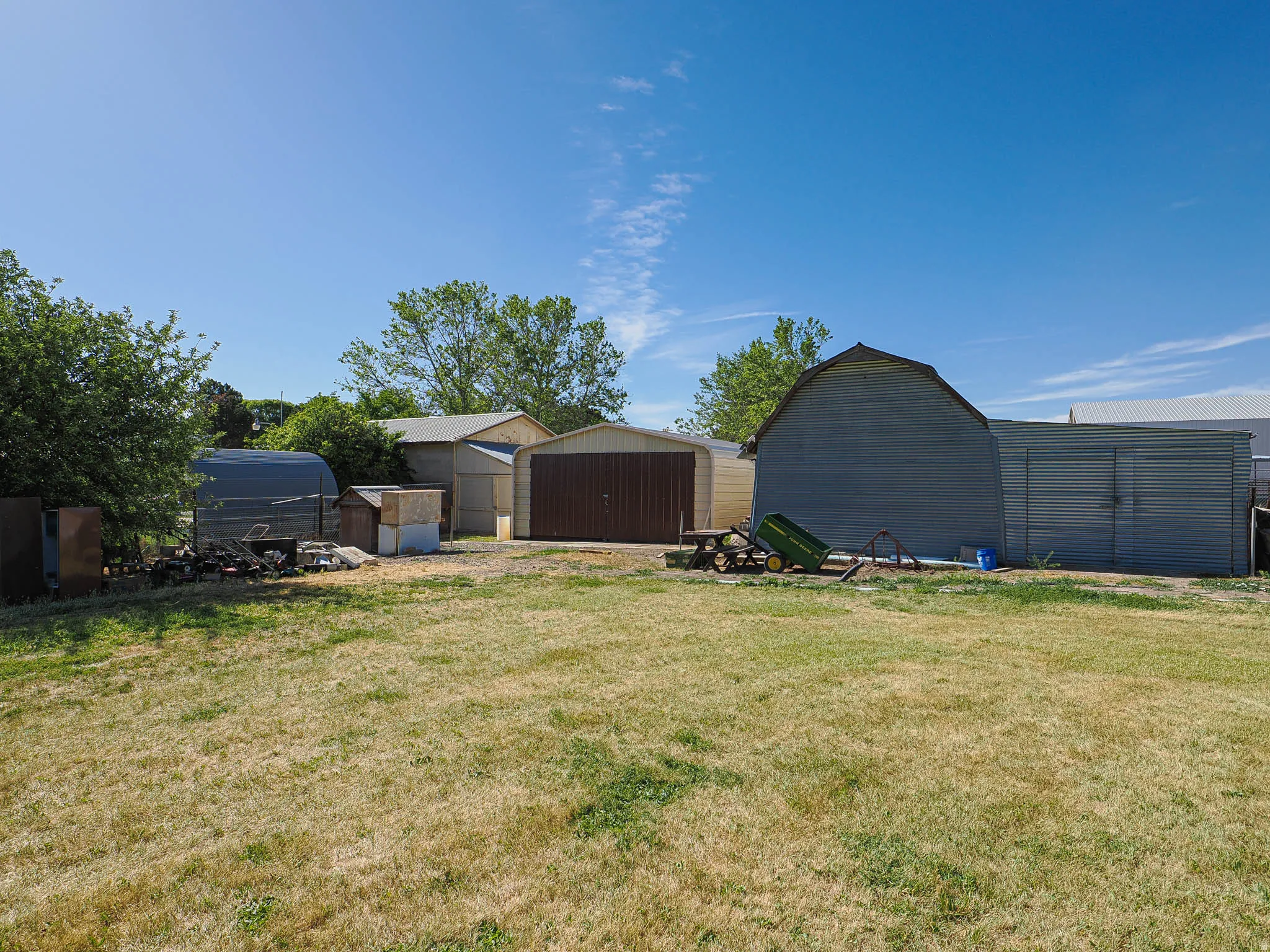 Expansive yard featuring multiple detached structures, including a large gray barn-style building, a gray metal shed with double doors, a brown garage with double doors, and additional storage sheds