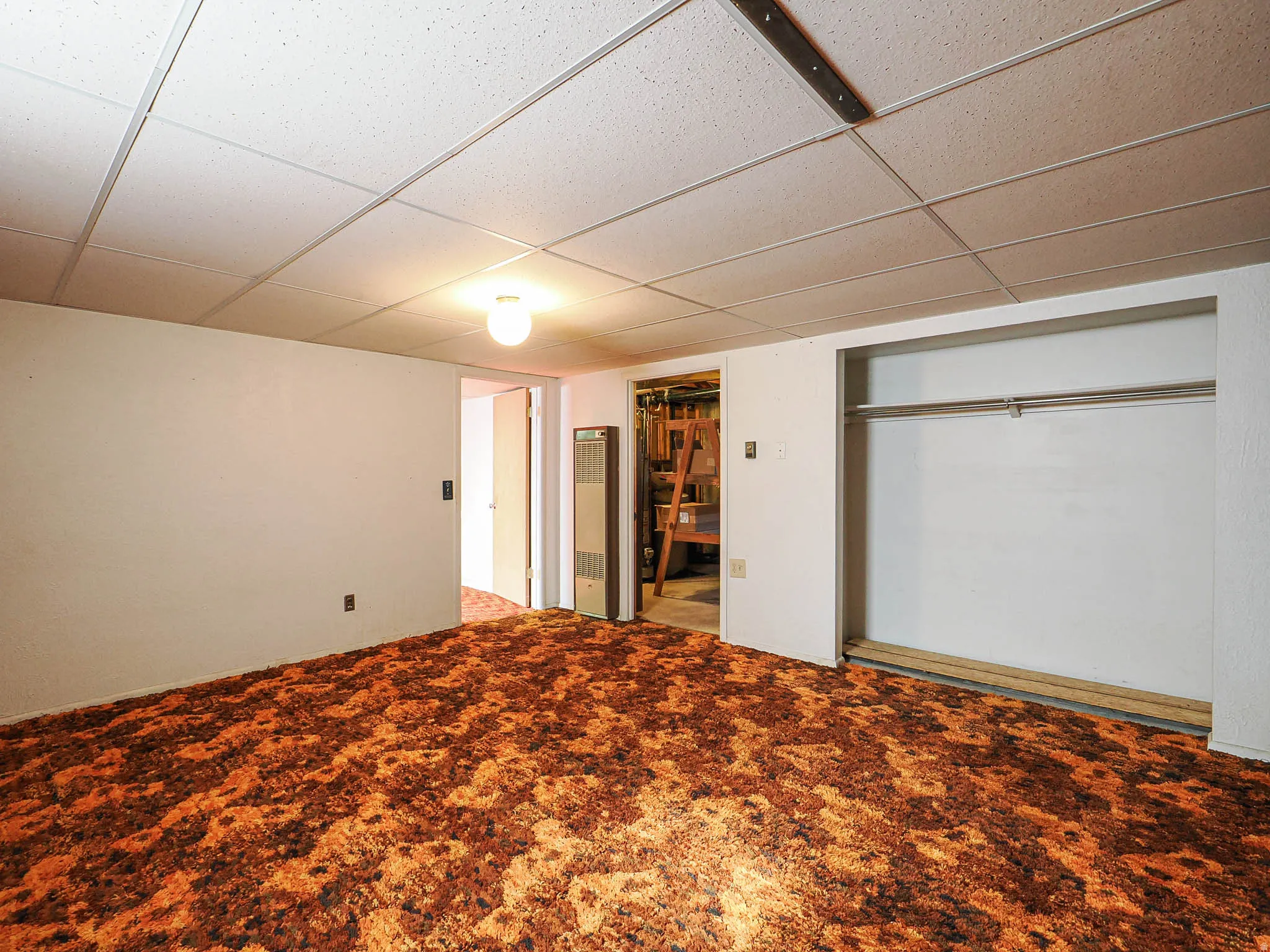 Room featuring wall-to-wall carpeting, drop ceiling with integrated lighting, a built-in closet with hanging rod, and a wall-mounted heating unit