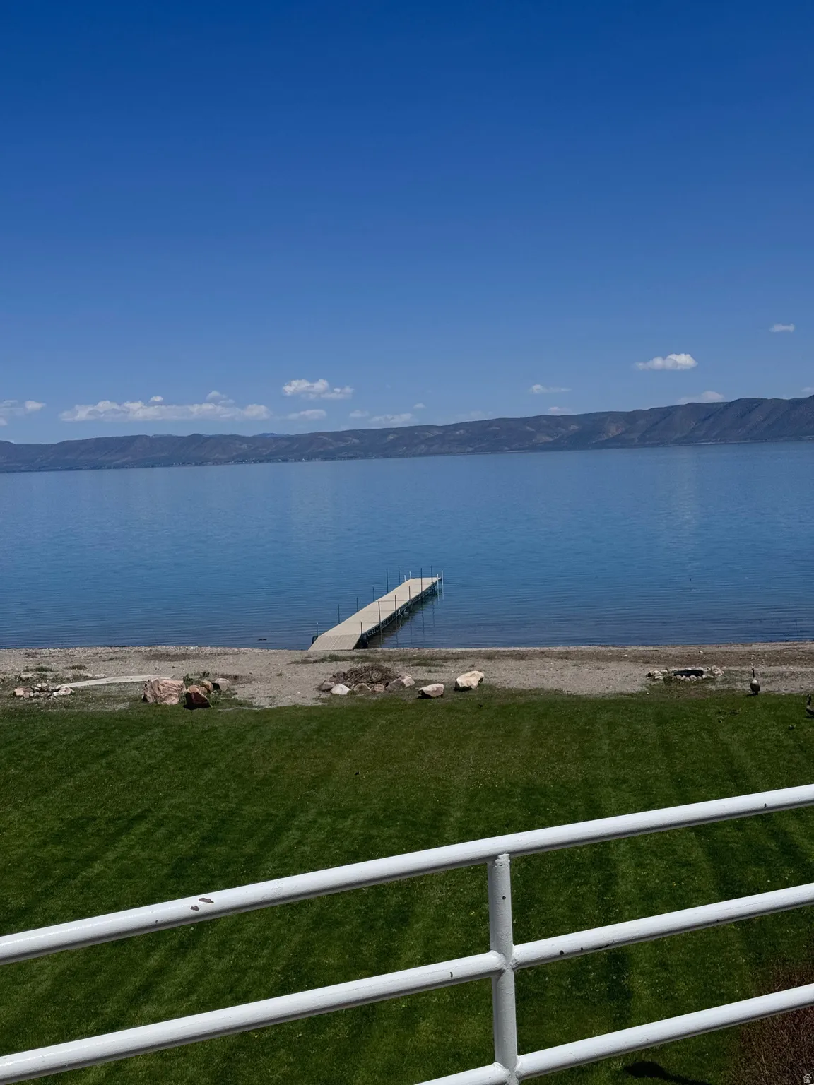 Expansive waterfront property featuring a private dock, a manicured lawn, a gravel beach, and distant mountain scenery