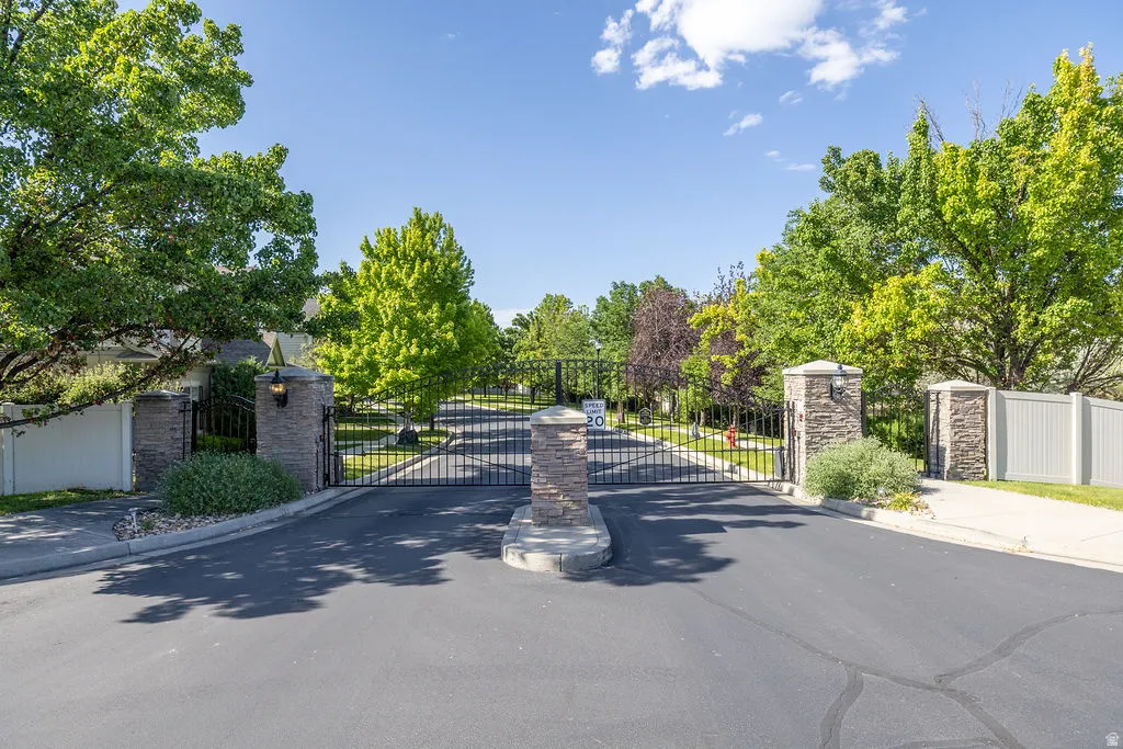 Gated community entrance featuring stone-clad pillars, wrought iron gates, mature landscaping, and an asphalt driveway