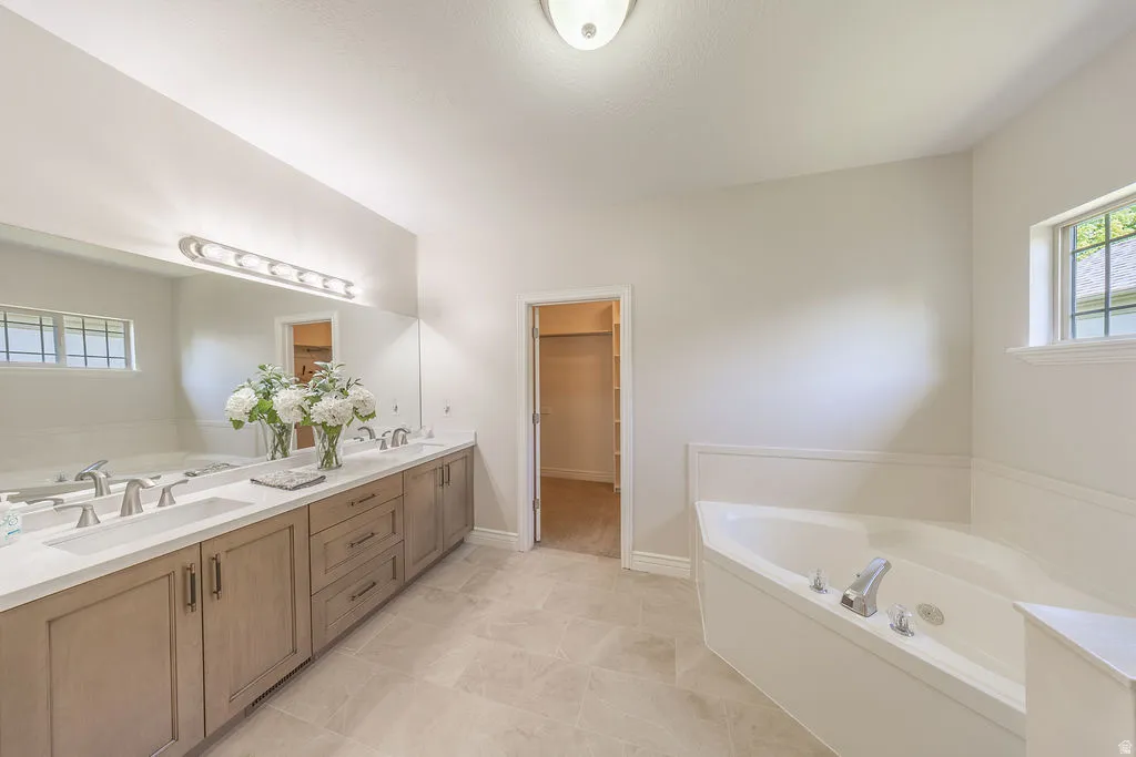 Spacious bathroom featuring a dual vanity with light wood-finish cabinetry and white countertops, a large mirror with a vanity light fixture, a corner soaking tub, and neutral-toned floor tiling