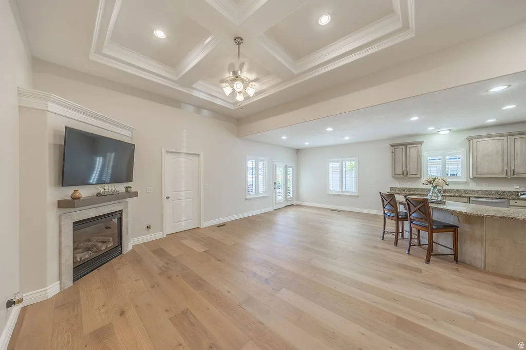 Open-concept great room featuring a coffered ceiling with recessed lighting, an electric fireplace with a stone surround, light wood-finish flooring, and a kitchen island with a granite-style countertop