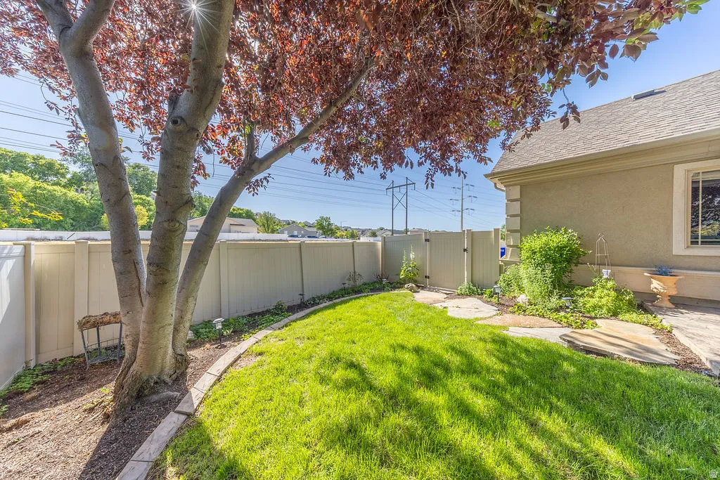 Enclosed side yard featuring a mature tree with red foliage, a manicured lawn, a gated privacy fence, stone pathway, and stucco exterior with decorative trim