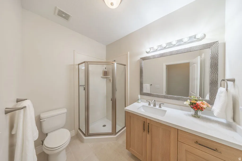Bathroom featuring a glass-enclosed corner shower, a single vanity with a white countertop and natural wood-finish cabinetry, a rectangular framed mirror with overhead lighting, and light-toned floor tiling