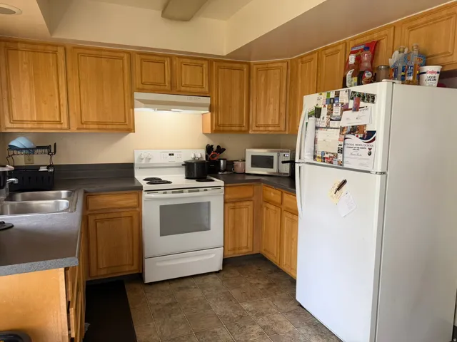 Kitchen featuring light wood cabinetry, white appliances, a double basin stainless steel sink, dark countertops, and tile flooring