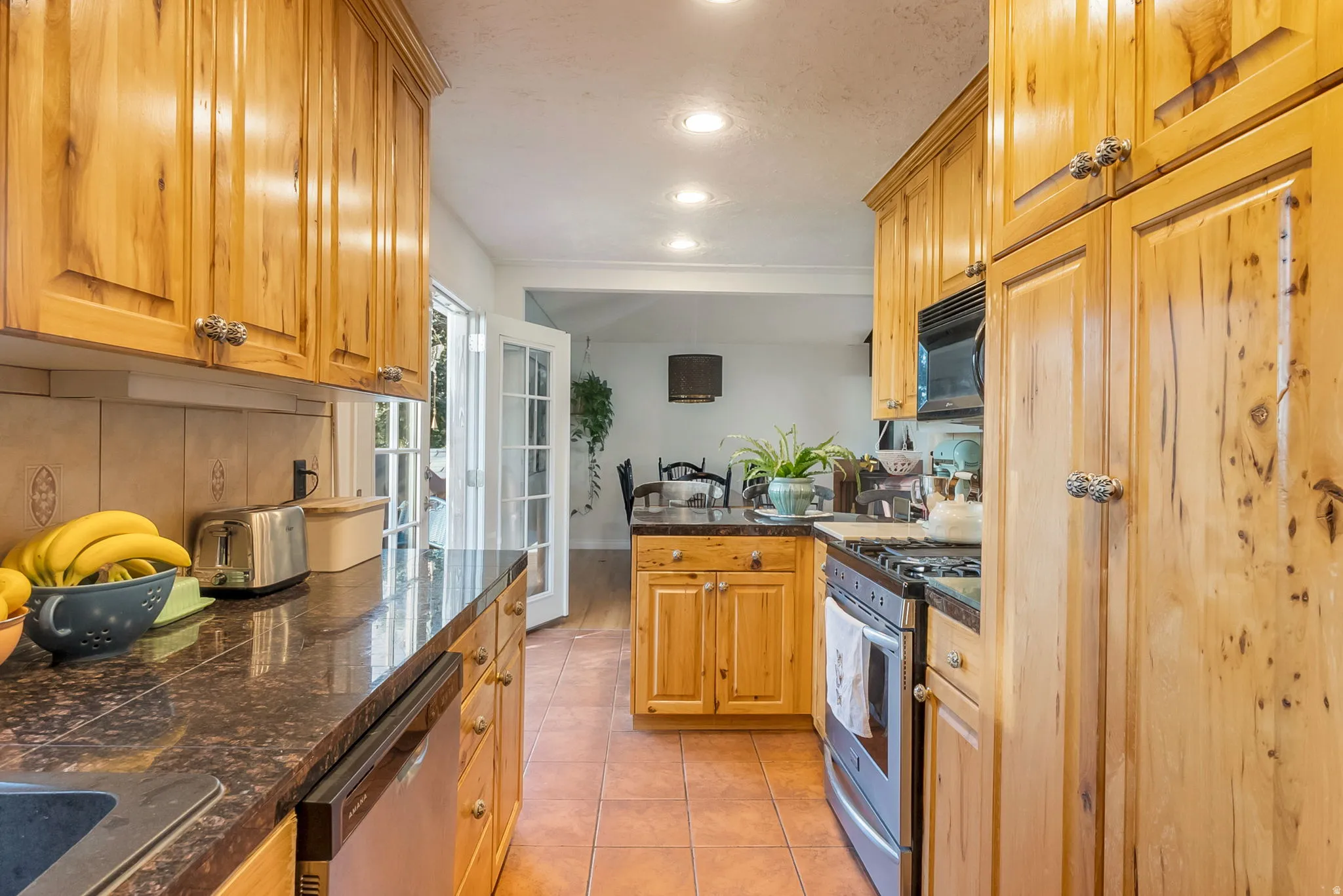 Kitchen featuring natural wood cabinetry, dark speckled countertops, stainless steel appliances, recessed lighting, and terracotta-toned floor tile