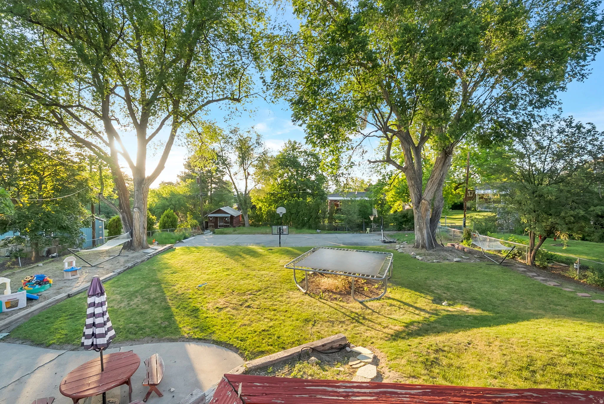 Spacious outdoor area with mature trees, a manicured lawn, paved patio, and a dedicated sport court