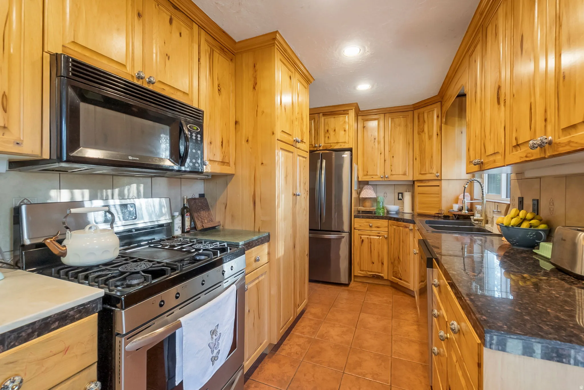 Galley kitchen featuring light wood cabinetry, dark stone countertops, stainless steel appliances, a tile backsplash, and ceramic tile flooring