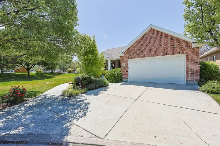 Brick facade residence featuring an attached garage with a white door, a concrete driveway, established landscaping, and mature trees