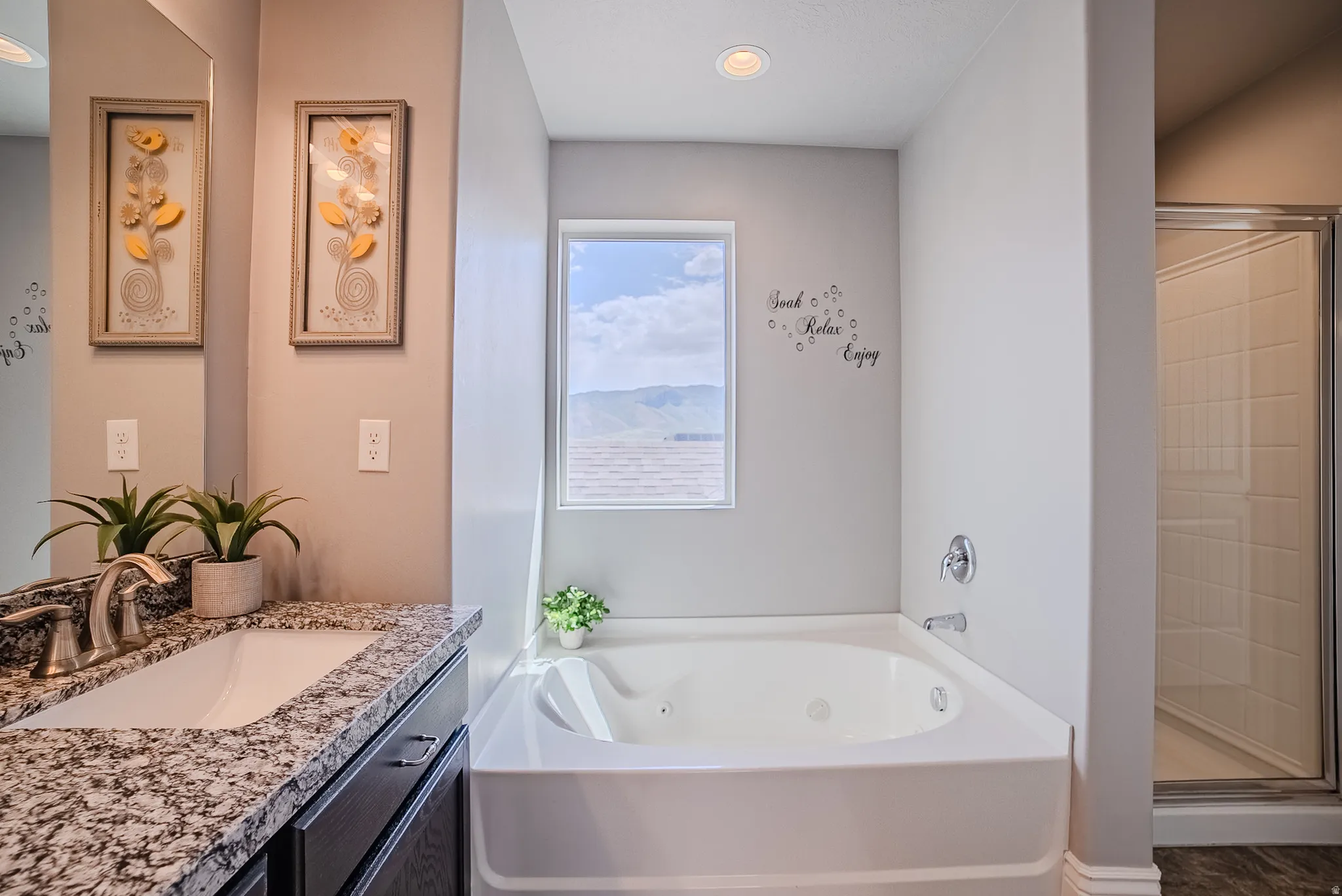 Bathroom featuring a white corner soaking tub, a window with mountain scenery, a granite-finish vanity with a single basin sink, a frameless mirror, and a glass-enclosed shower